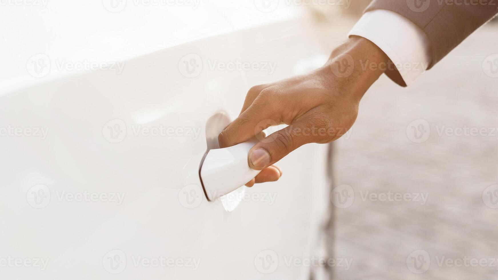 Entrepreneur hand is shown opening the door of a white car. The hand is reaching for the door handle and pulling it open. The car is parked in a sunny location. photo