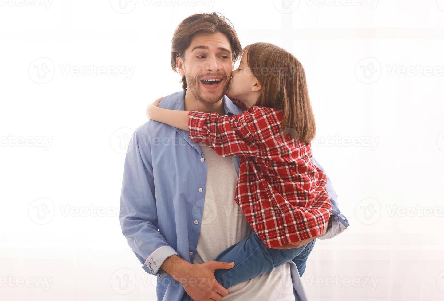 Handsome young dad happy to be kissed by little daughter, man holding preschool girl next to window at home, free space photo