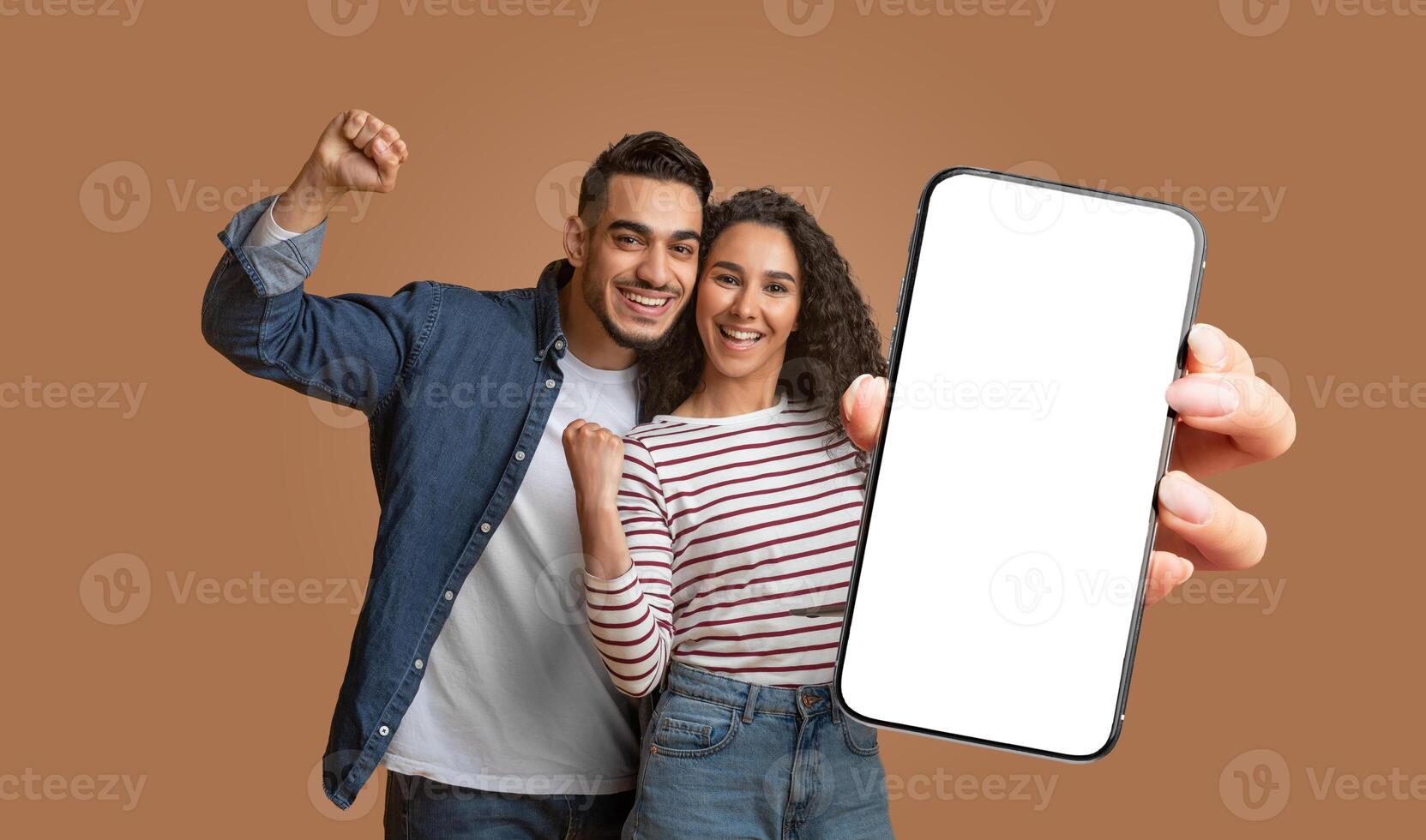 This image shows middle eastern couple standing in front of a brown background. They smiling and excited, with their fists raised in the air. The woman is holding a smartphone with a blank screen. photo