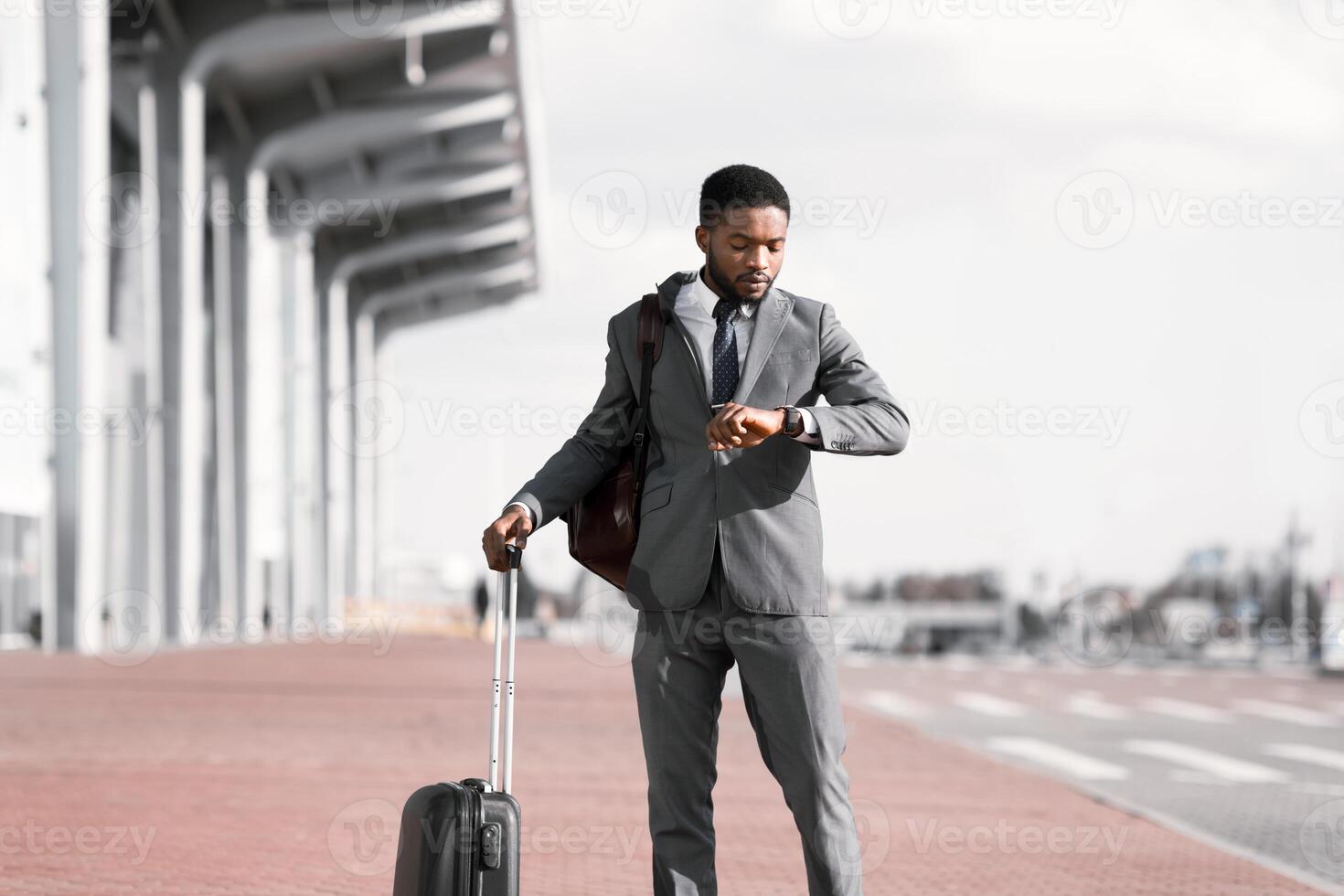 A professional black man with a suitcase is checking his watch outside an airport. The mood is efficient and on-the-go, with the modern airport setting creating a sense of urgency. photo