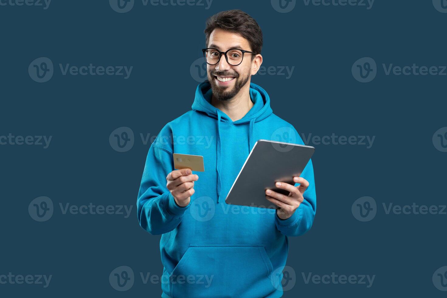 A cheerful man, dressed in a blue hoodie, holds a credit card in one hand and a tablet in the other. He appears excited about online shopping, showcasing the convenience of digital transactions photo