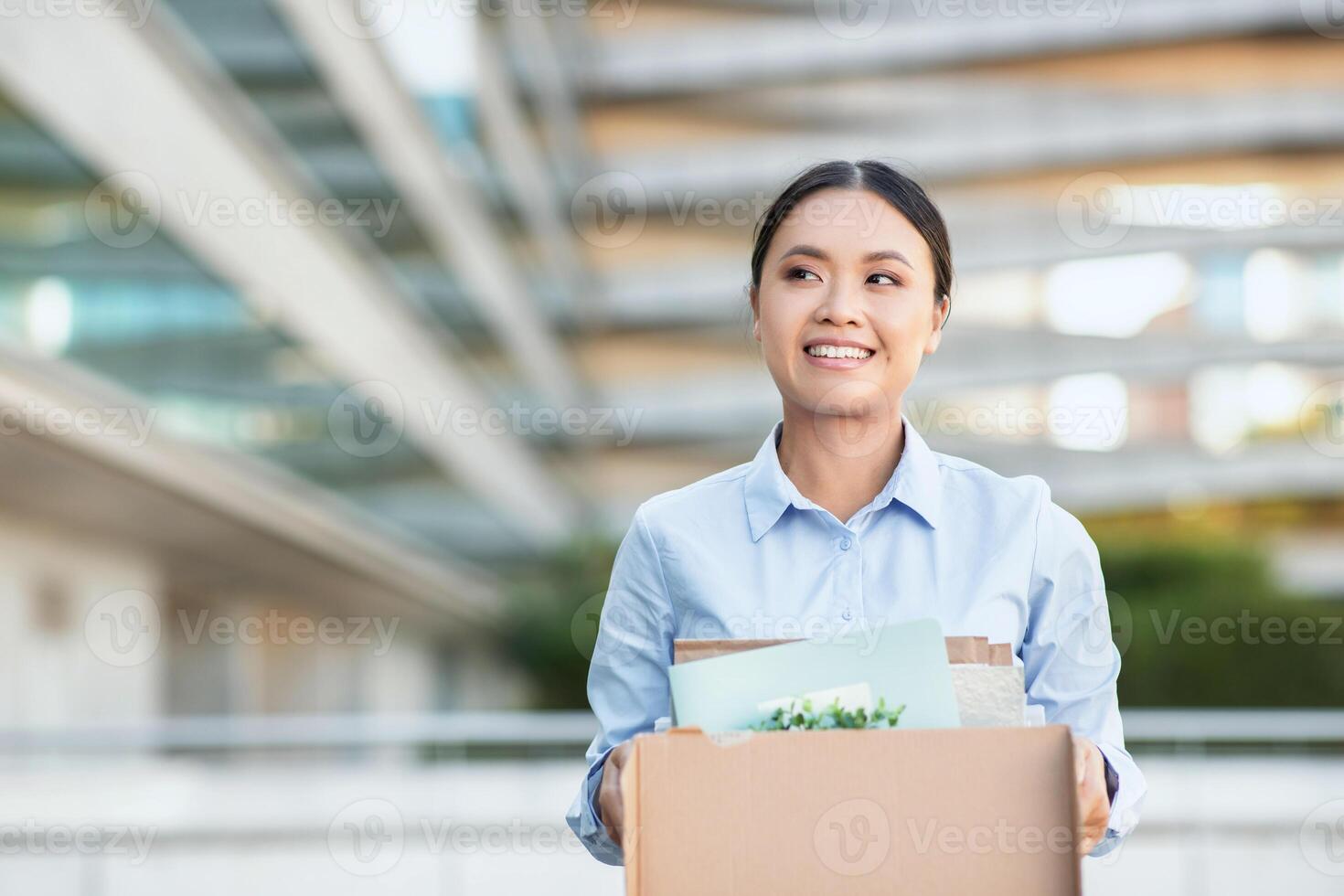 Excited Asian businesswoman is standing while holding a cardboard box. She appears focused and determined, possibly moving office or transporting items photo