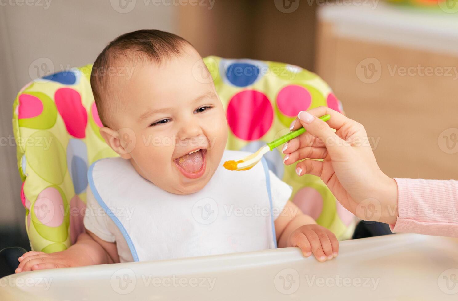 A joyful baby boy sits in a colorful high chair, eagerly opening their mouth as a caregiver feeds them with a spoon of baby food in a bright, cheerful kitchen. photo