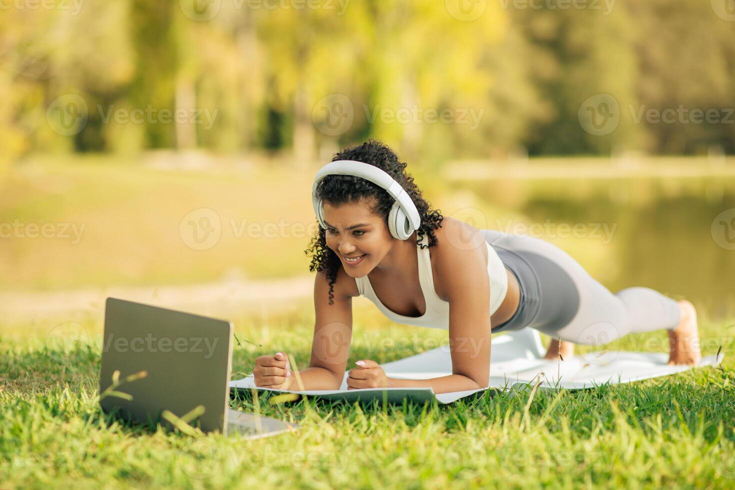 A woman in athletic wear is seen doing a plank exercise on a yoga mat while watching a laptop, with headphones on in a sunny park. photo