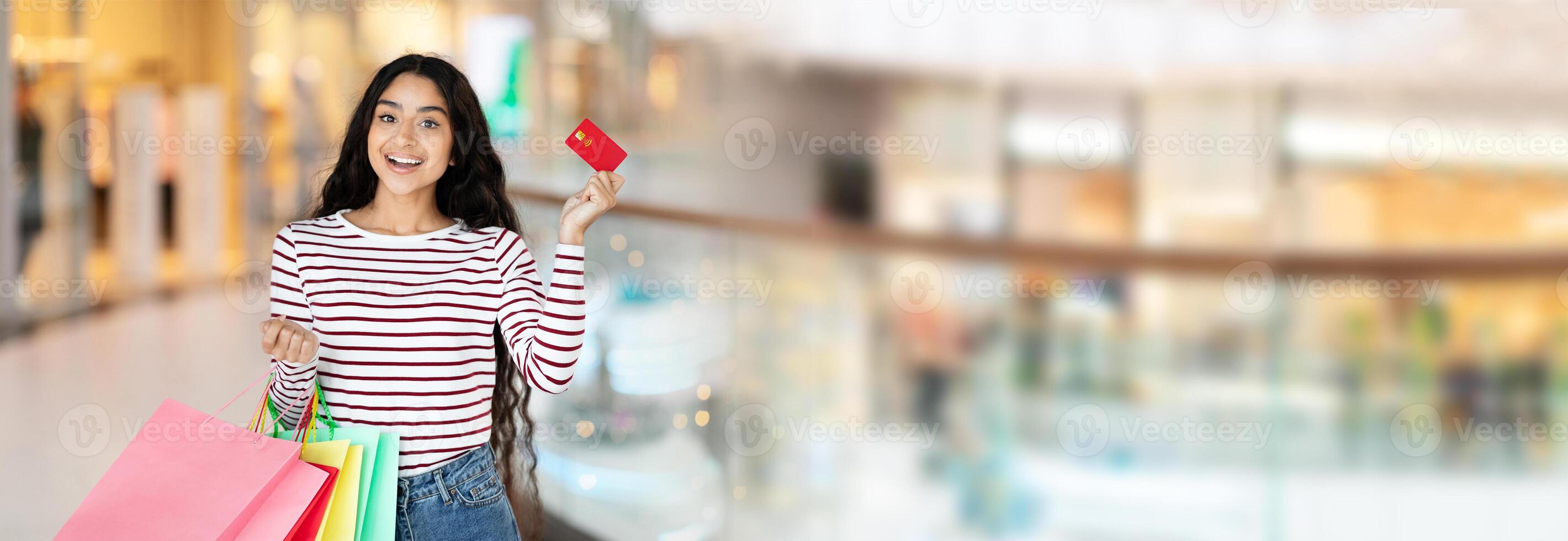 Eastern young woman with long, dark hair is standing in a modern shopping mall. She is smiling happily while holding a red credit card and colorful shopping bags, copy space photo