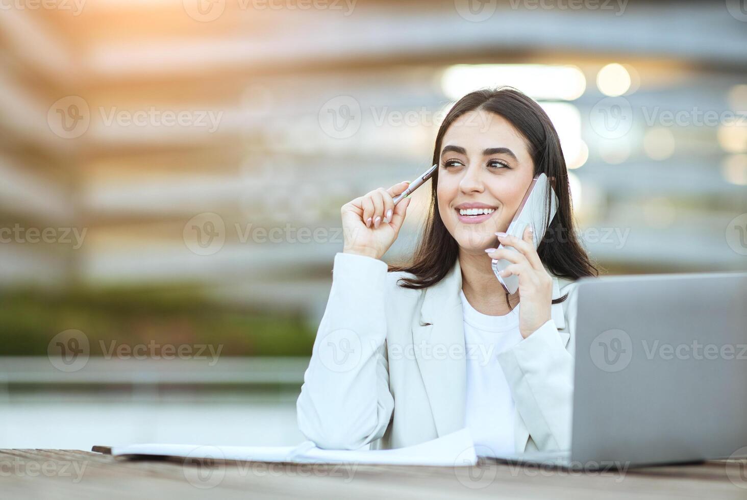 A smiling young businesswoman in a white blazer is sitting outside an office building. She is working on her laptop and talking on her cell phone. There are papers in front of her on a wooden table. photo