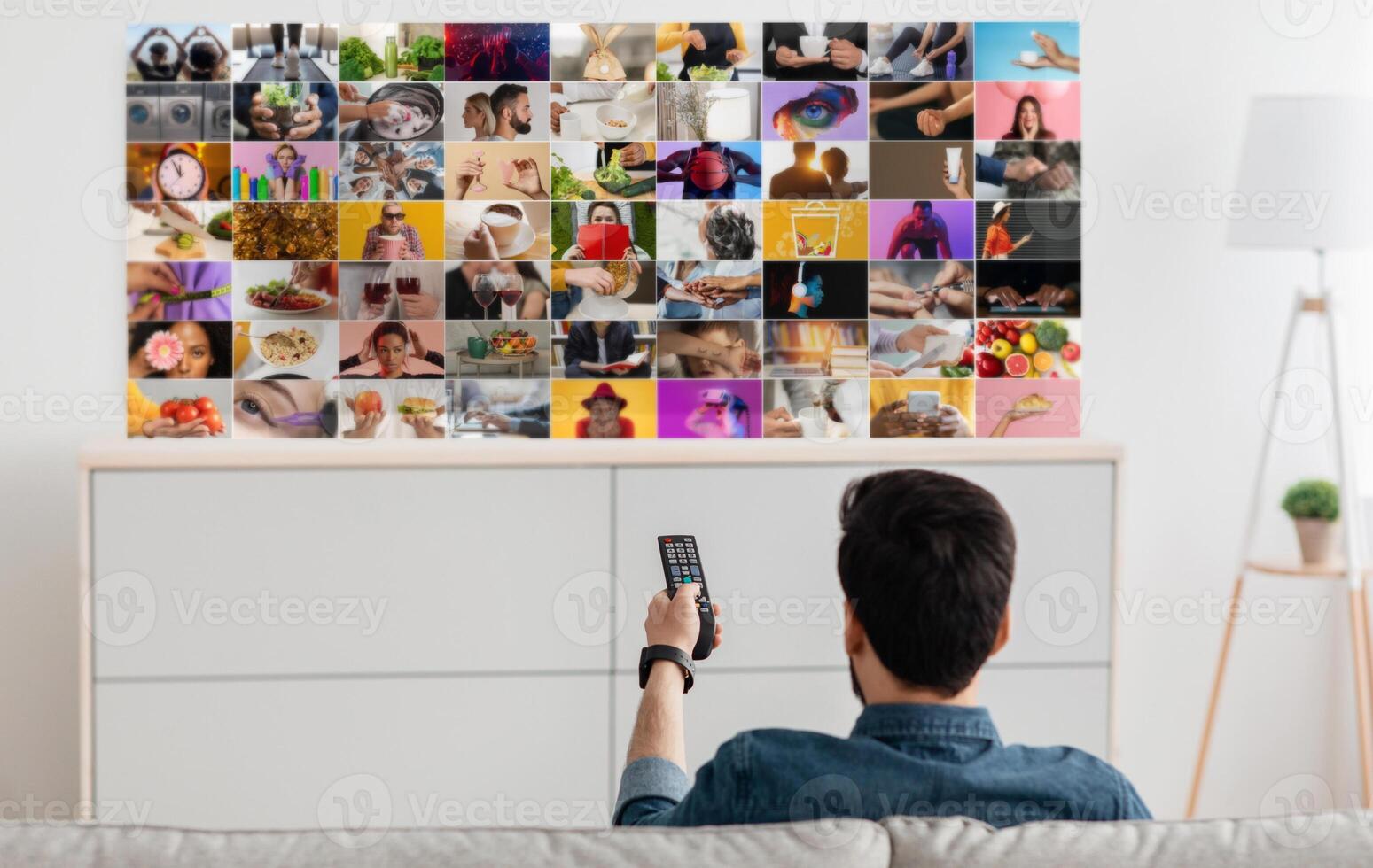 A man sits on a couch in a living room, facing a television screen. He holds a black remote control in his right hand and is using it to navigate the televisions content. photo