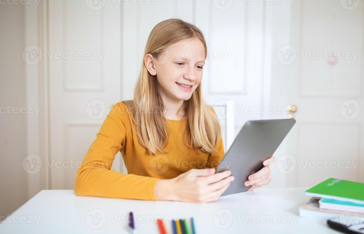 A teen girl with long blonde hair sits at a white desk in a home office, using a tablet computer. She is smiling and looking at the screen. A stack of books and some colored pencils are on the desk. photo