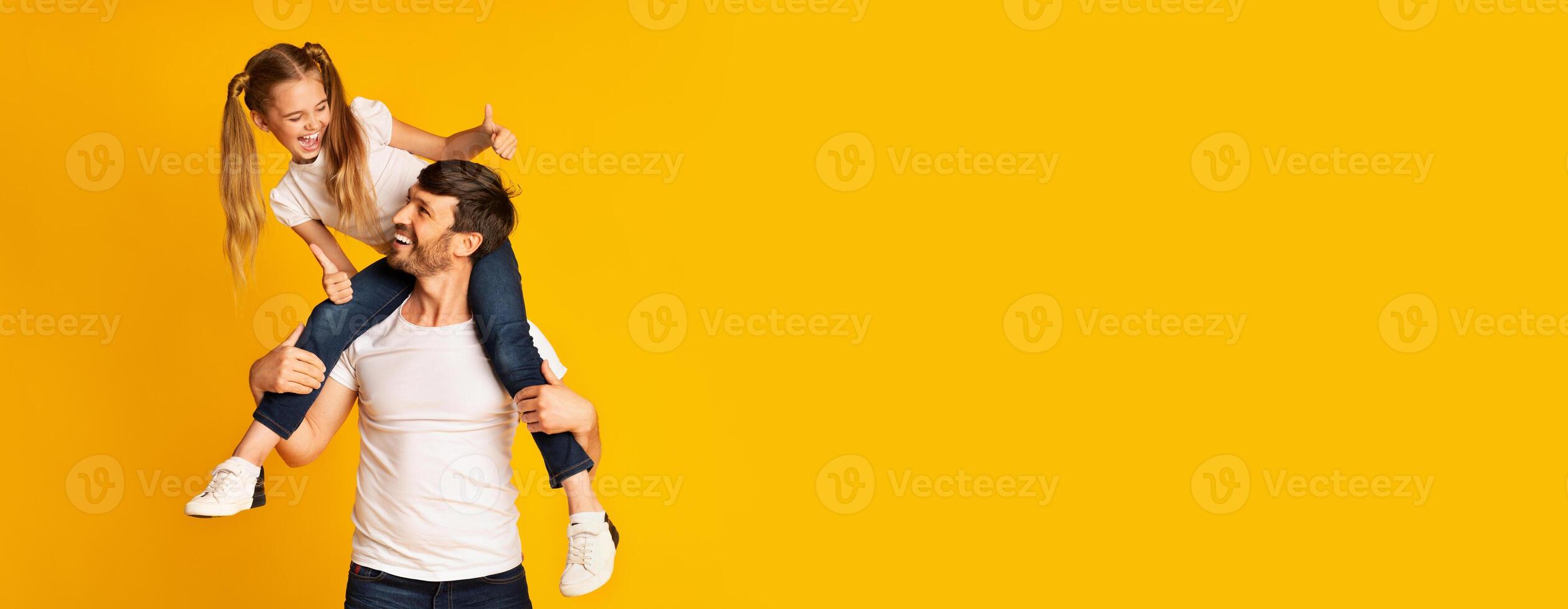 A father carries his daughter on his shoulders, both smiling happily against a vibrant yellow background. The playful interaction showcases their joyful bond and lighthearted spirit, copy space photo
