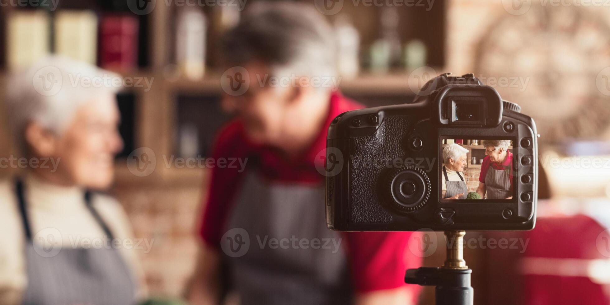A happy senior couple, wearing aprons, stand in a home kitchen and film a cooking . The couple smiles as they look at the camera, which is mounted on a tripod. photo