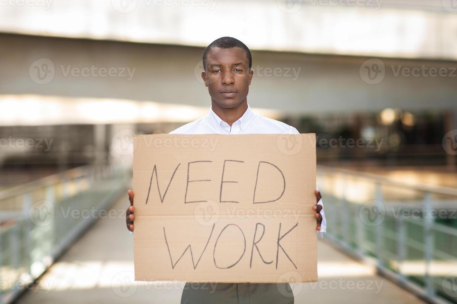 A young African American man stands in an urban area, looking serious as he holds a large cardboard sign that clearly states he needs work. photo