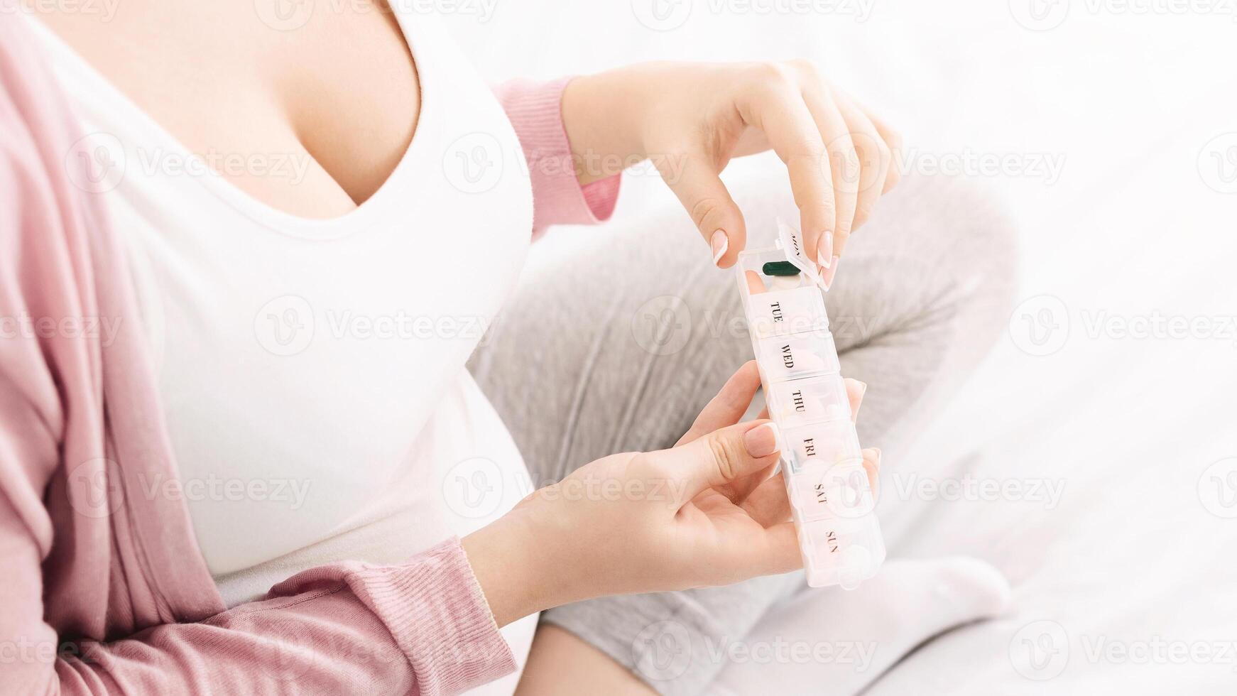A woman is organizing her daily medication into a pillbox while sitting comfortably on a bed. She is wearing casual, home attire, with a soft focus on her hands and the pillbox, cropped photo