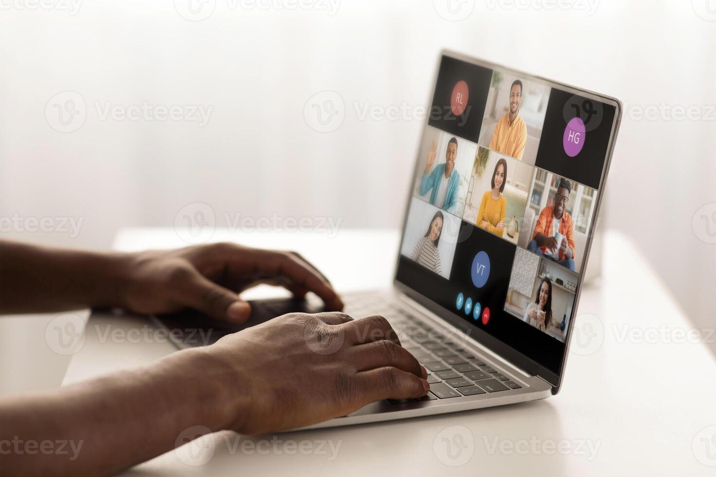 A person is sitting at a desk, using a laptop to participate in a conference with several other people. The laptop screen displays a grid of windows, each showing a different participant photo