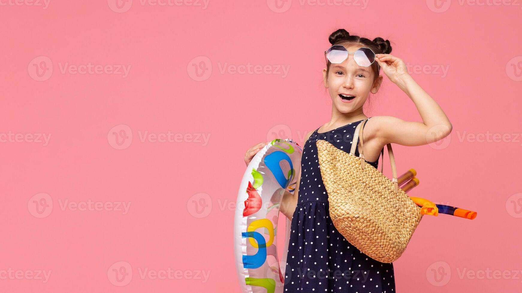 A young girl with her hair in two buns holds a colorful swim ring and a straw bag, ready for a fun-filled summer day, copy space photo