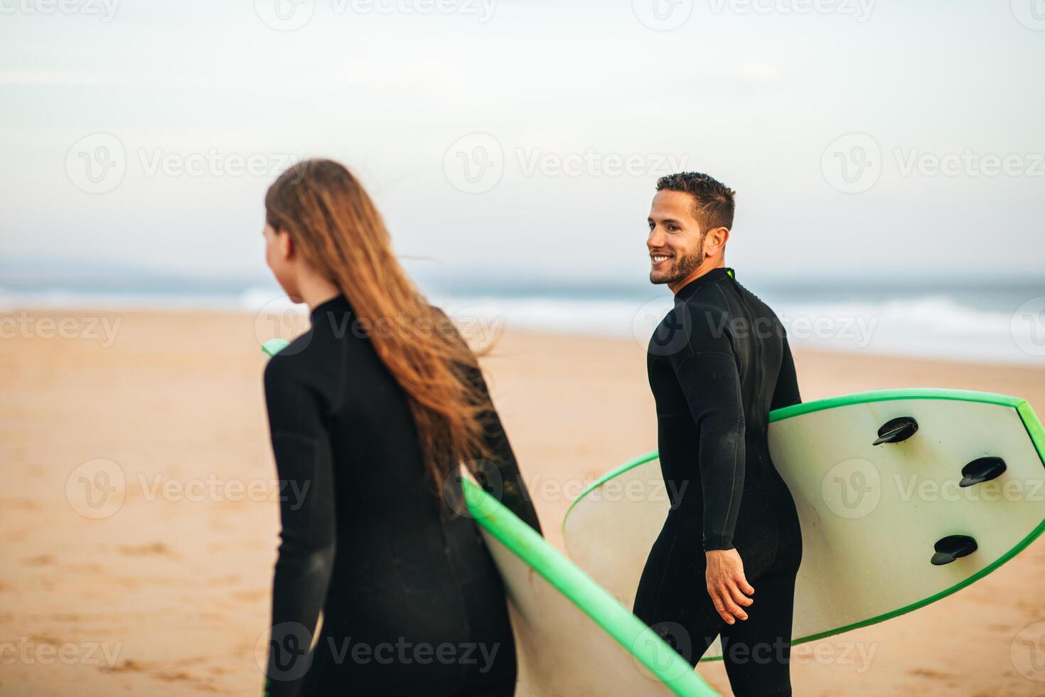 A young couple in wetsuits walk along a sandy beach with their surfboards, heading towards the water. The man is smiling and looking back at the camera, while the woman looks forward. photo