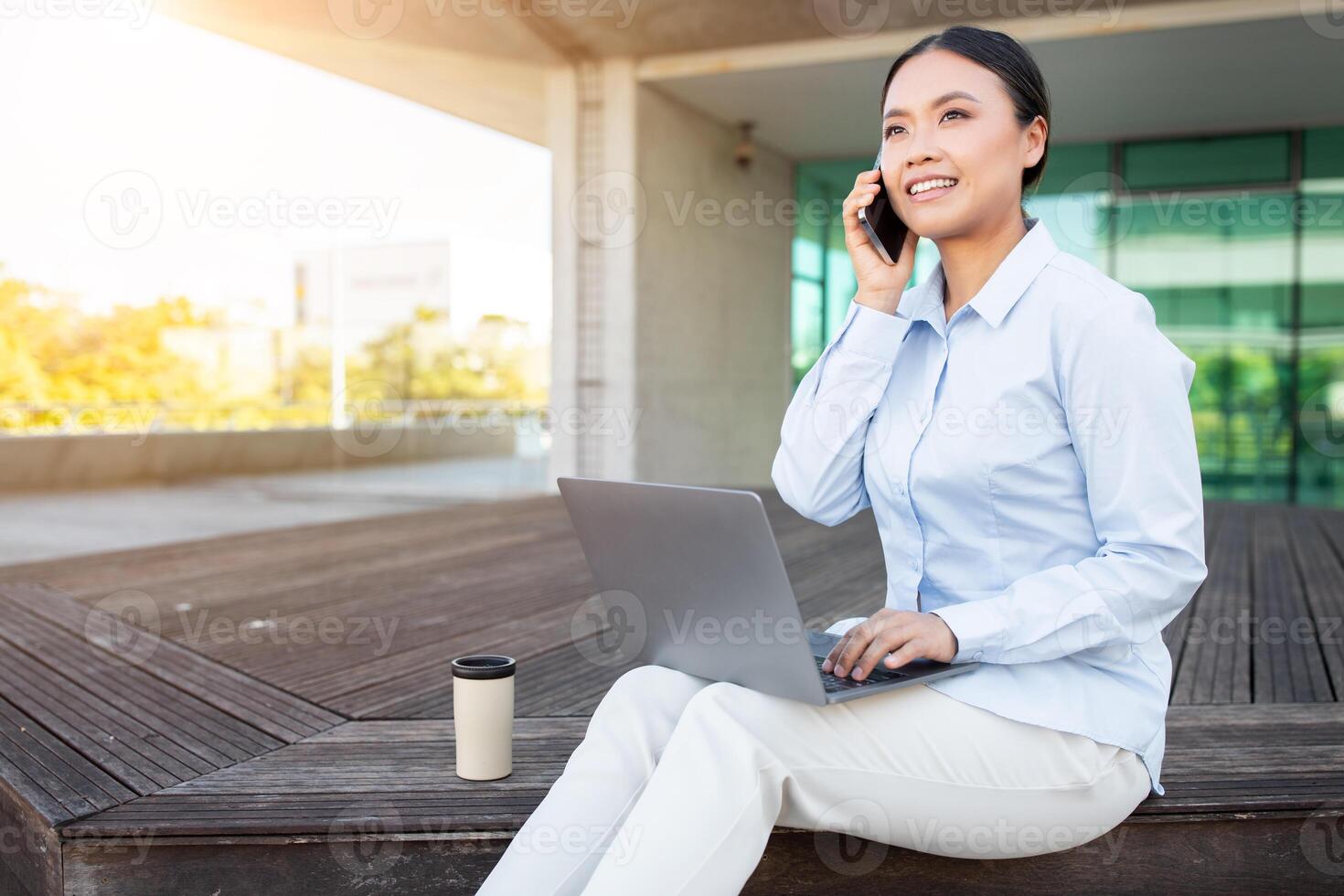 Asian woman sits comfortably on a wooden platform, working on her laptop while speaking on her phone. The bright afternoon sunlight illuminates her focused expression, copy space photo