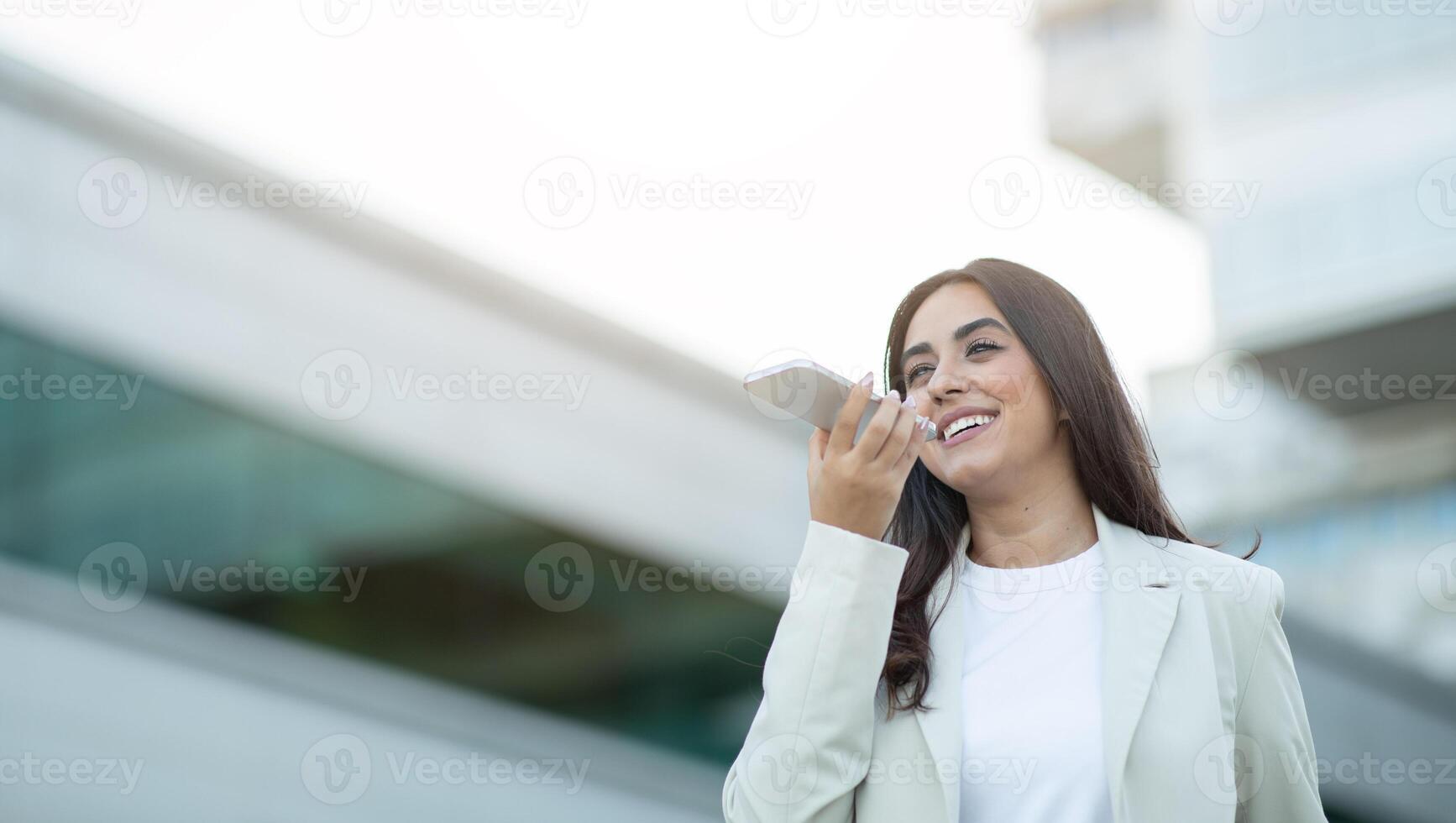 A young woman in a white blazer smiles as she uses the voice recognition feature on her smartphone. She stands outside in an urban setting, with buildings in the background, copy space photo