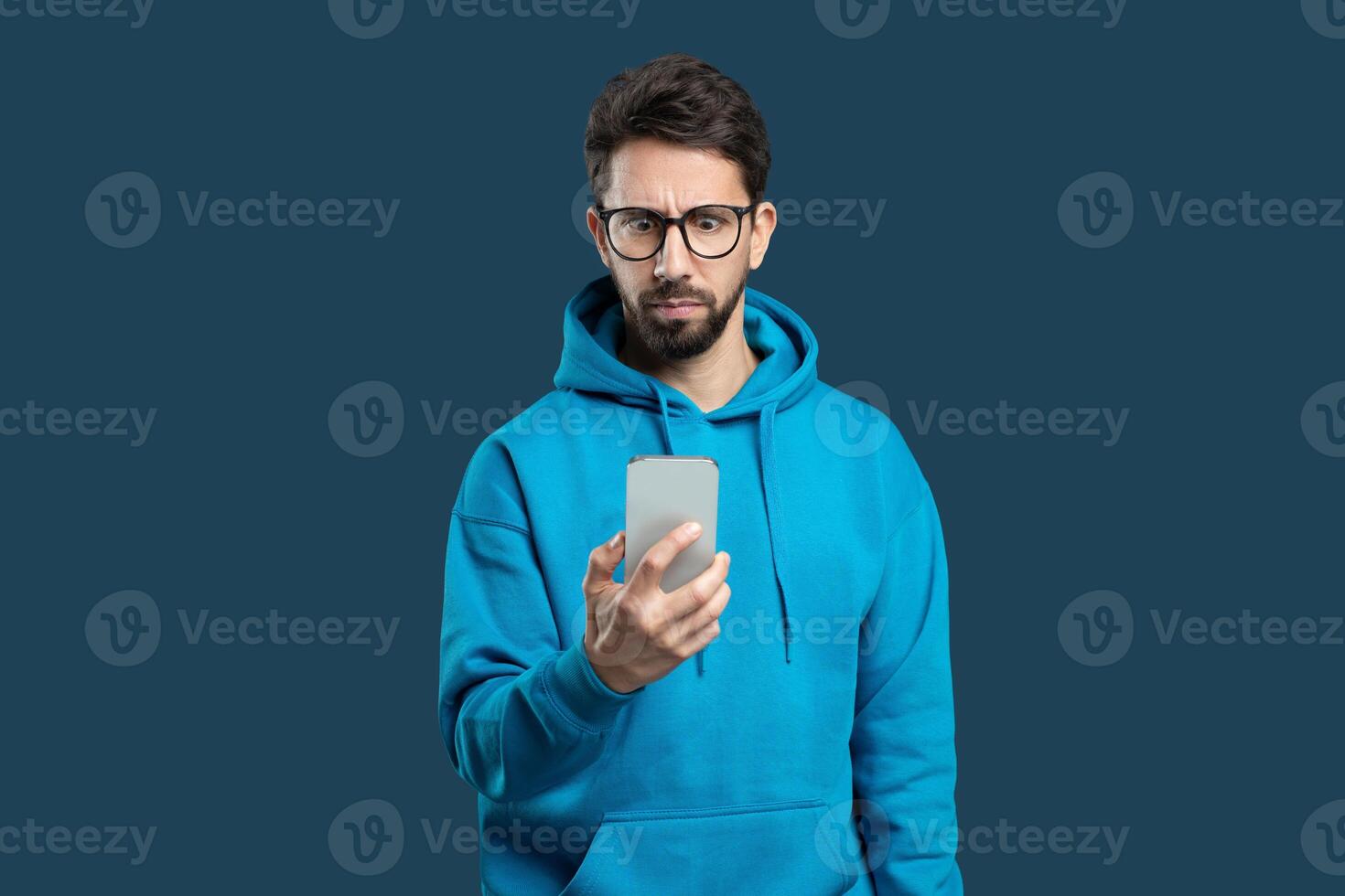 A young man wearing a blue hoodie is holding a smartphone in one hand while sporting a serious expression. He appears to be engaged in a focused activity, possibly examining content on the device photo