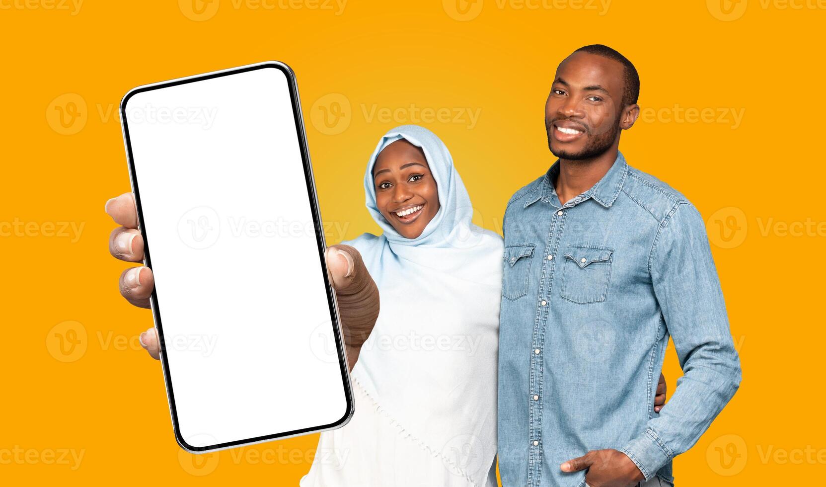Black woman in a light blue hijab smiles and points to a blank phone screen while standing next to a man in a denim shirt. Both are looking directly at the camera. photo