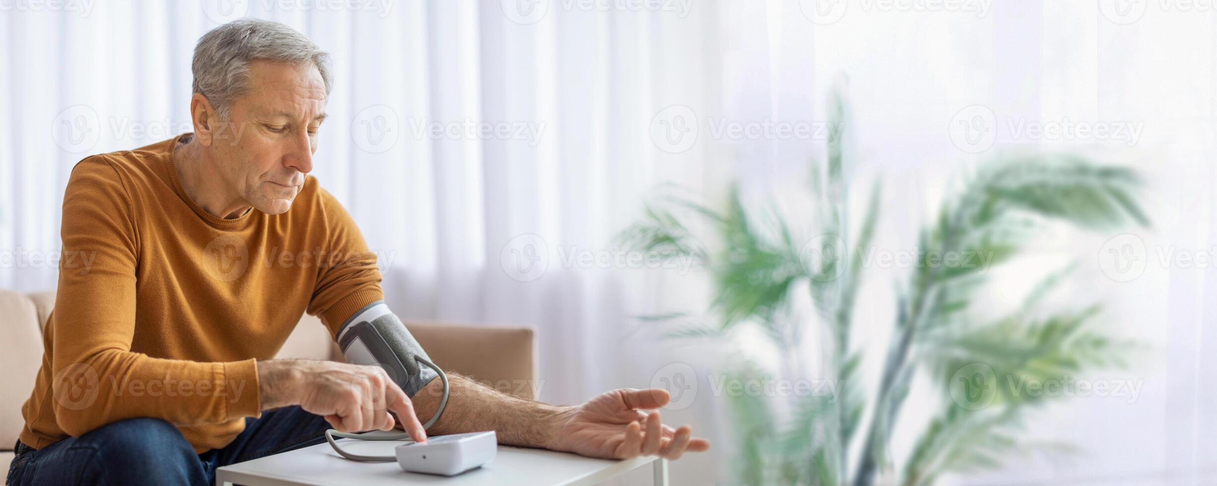 An elderly man wearing a yellow sweater sits at a table in his living room and uses a digital blood pressure monitor to check his blood pressure, copy space photo