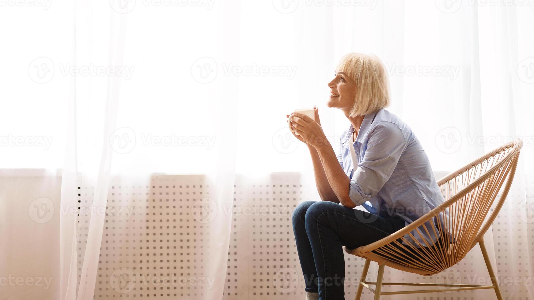 Senior woman sitting on chair in front of window and drinking tea, side view, copy space photo