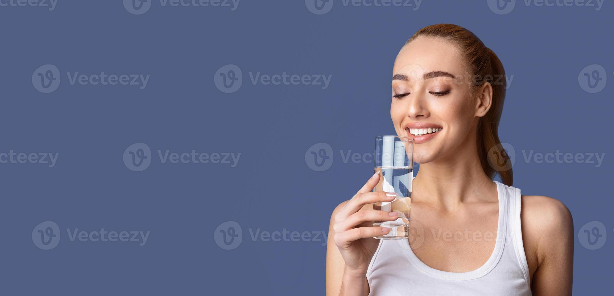 A woman in a white tank top is drinking water from a glass while looking at the water with her eyes closed and a smile on her face. Her hair is pulled back, standing against a blue background. photo