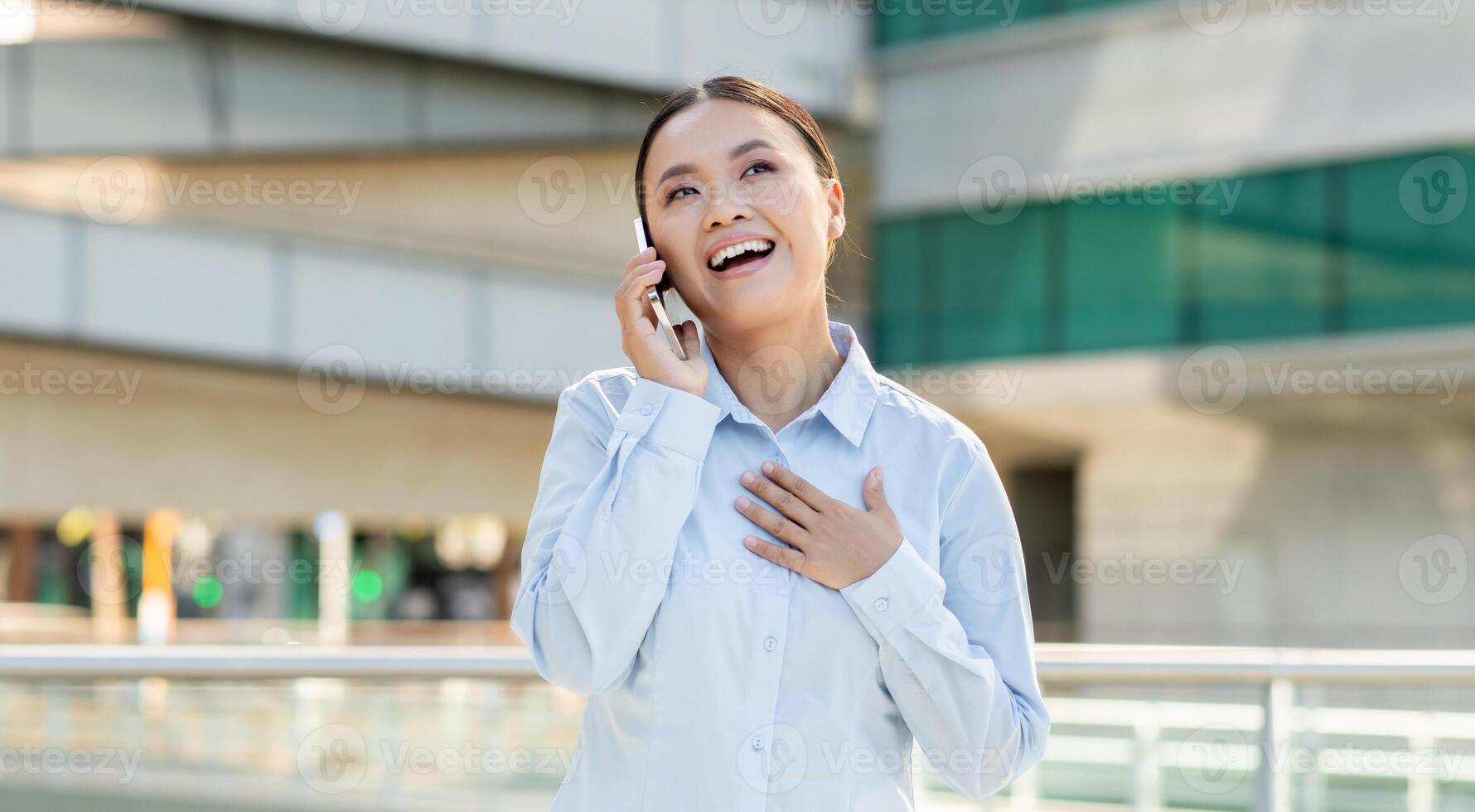 Asian young woman stands in an urban setting, smiling as she speaks on her mobile phone. Her genuine expression reflects happiness as she interacts with someone, possibly sharing exciting news photo