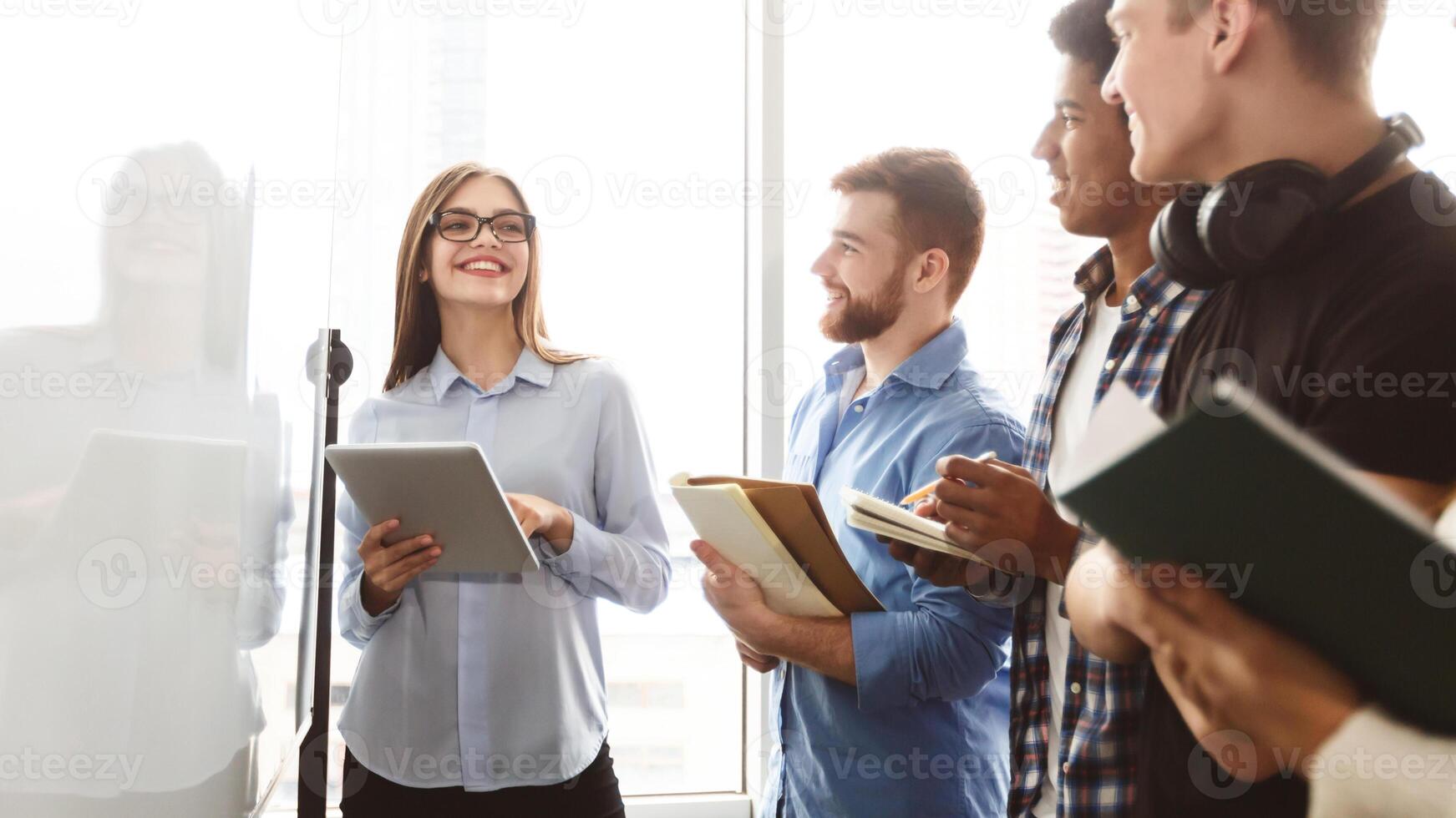 A diverse group of multiracial young people standing in front of a whiteboard, engaged in a discussion or presentation. Some are pointing at the board while others are taking notes photo