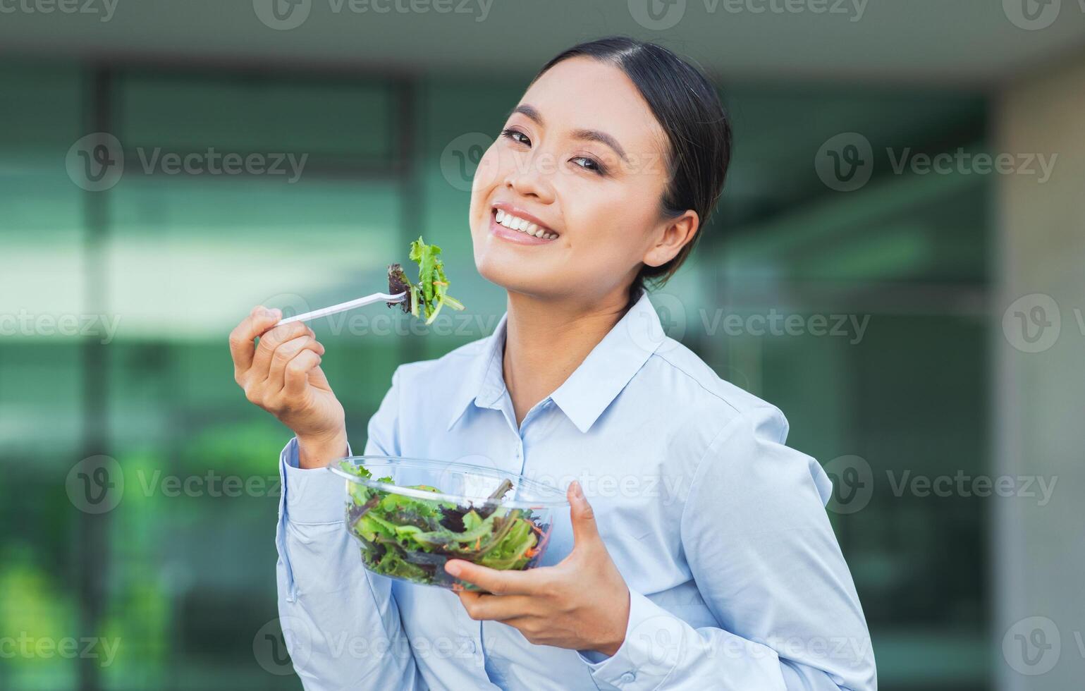 Chinese woman is sitting at a table, holding a fork and eating a bowl of freshly made salad. She appears focused on her meal, taking bites and chewing thoughtfully. photo