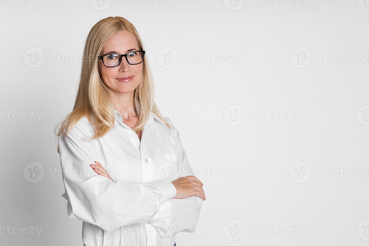 A blonde woman wearing glasses stands with her arms crossed in front of a white background. She is wearing a white shirt and looks directly at the camera with a slight smile on her face, copy space photo