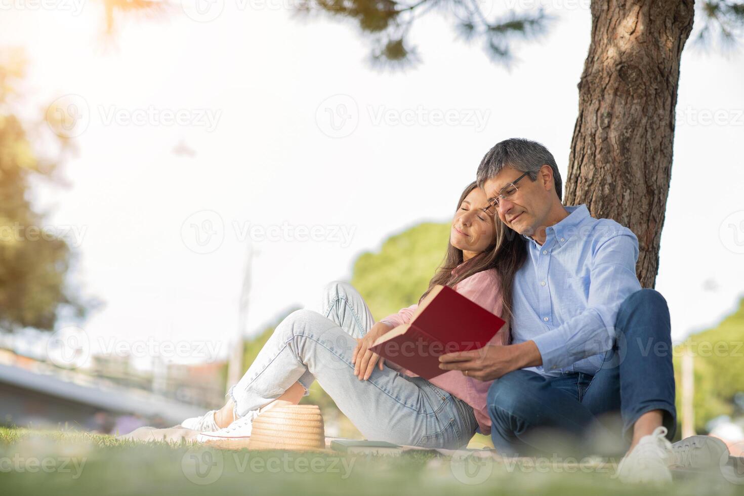A couple enjoys a cozy moment under a tree, sharing a book in a sunny park. Their relaxed posture and warm expressions reflect a peaceful spring afternoon together. photo