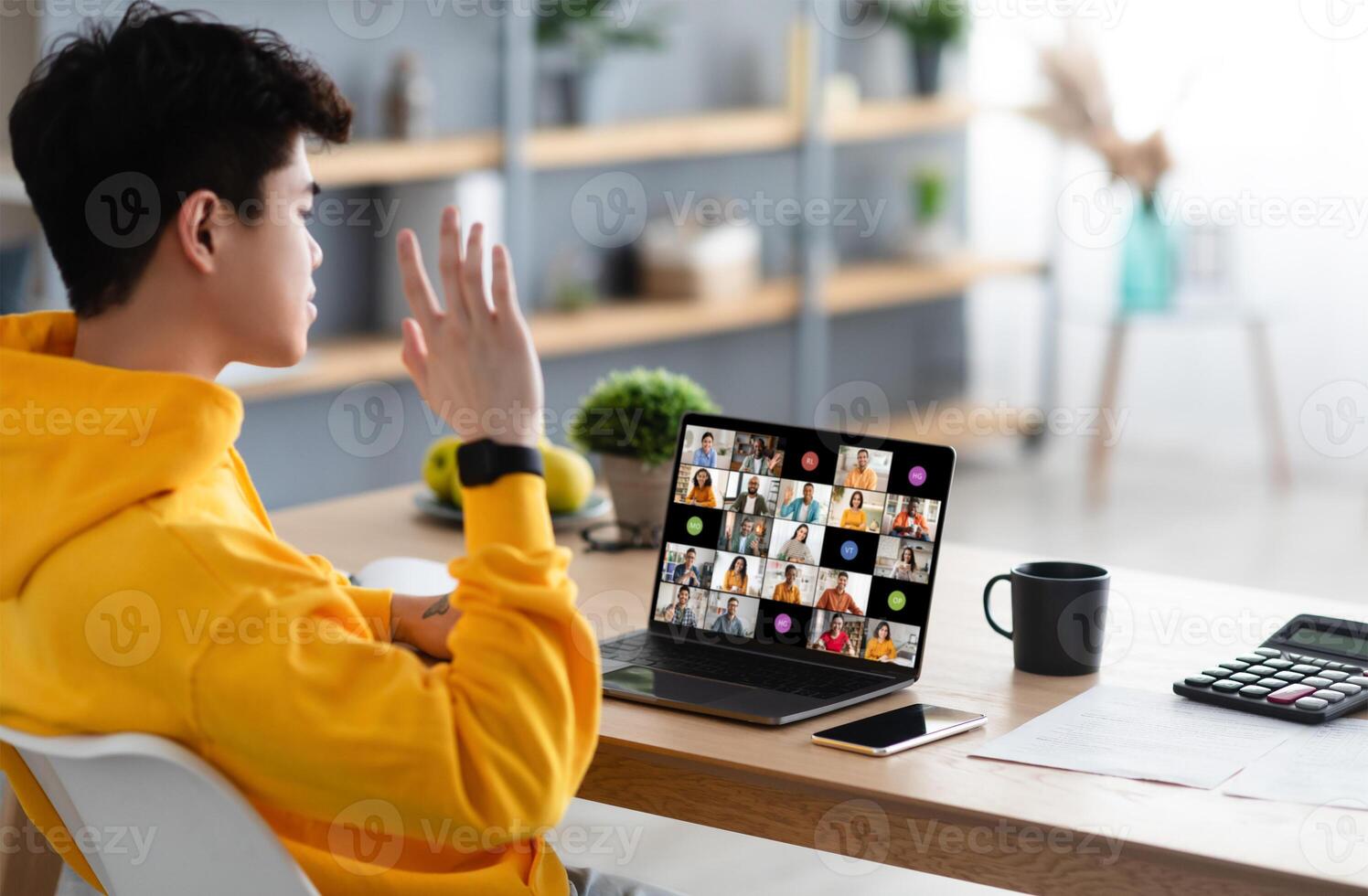 A young man sits at a desk at home, looking at his laptop screen. He is participating in a conference with many other people on the screen. His hand is raised photo