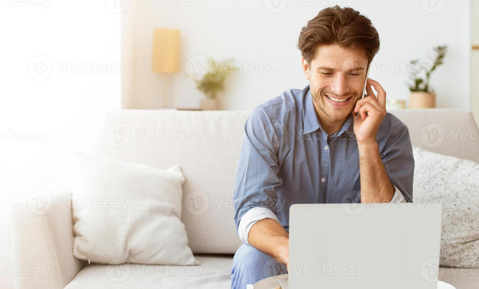 A man smiles while sitting on a couch, using a laptop and talking on the phone. He appears to be relaxed and happy. photo