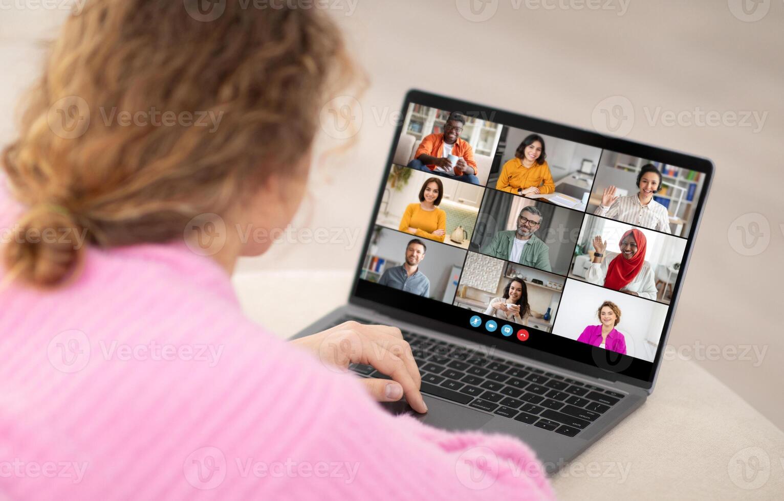A woman is sitting at a desk and looking at a laptop screen. She is participating in a conference call with eight other people. The people on the screen are in a grid layout photo