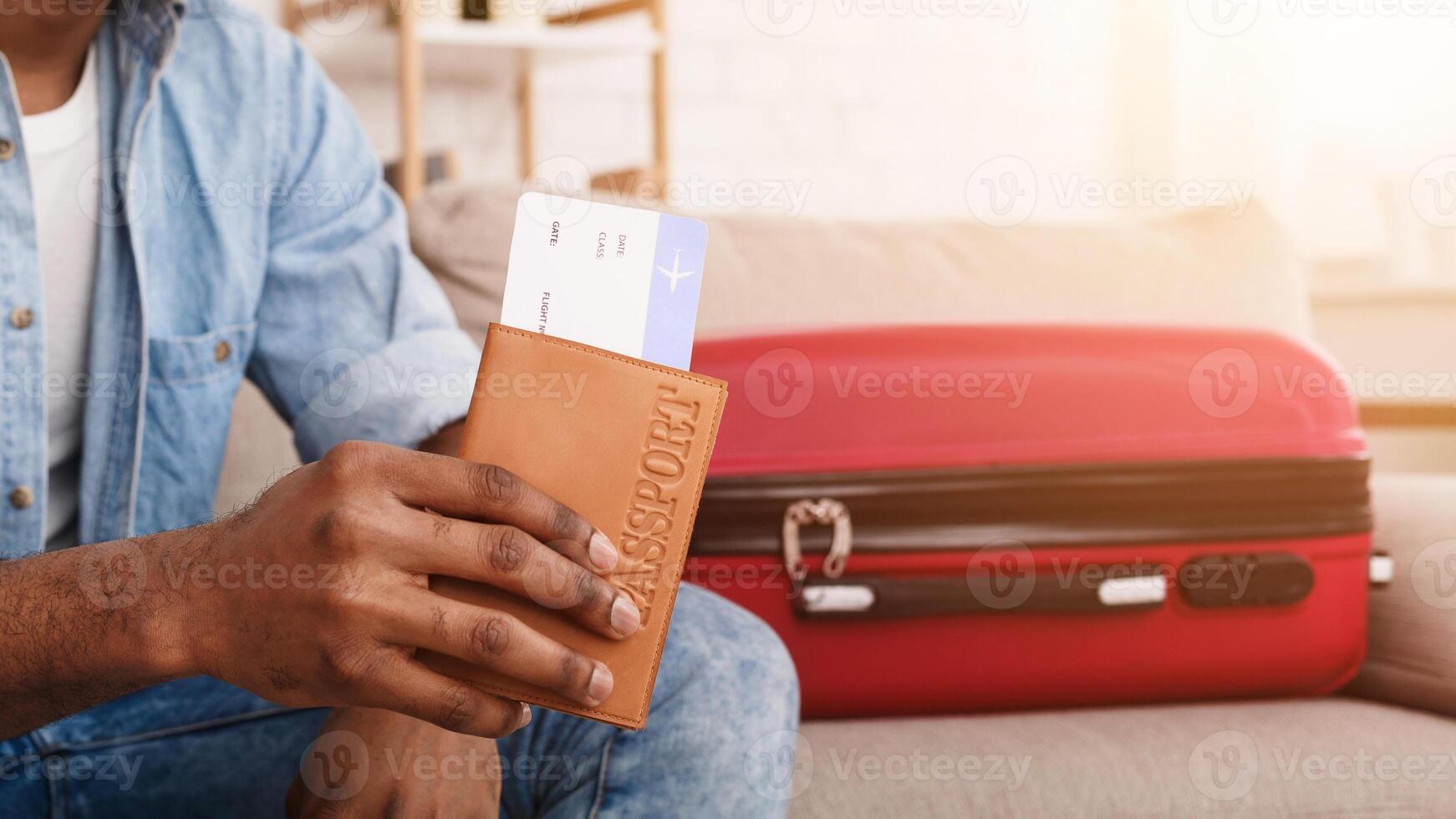 Cropped of black man is seated, displaying a passport and boarding pass, suggesting he is prepared for an upcoming journey. A red suitcase next to him photo