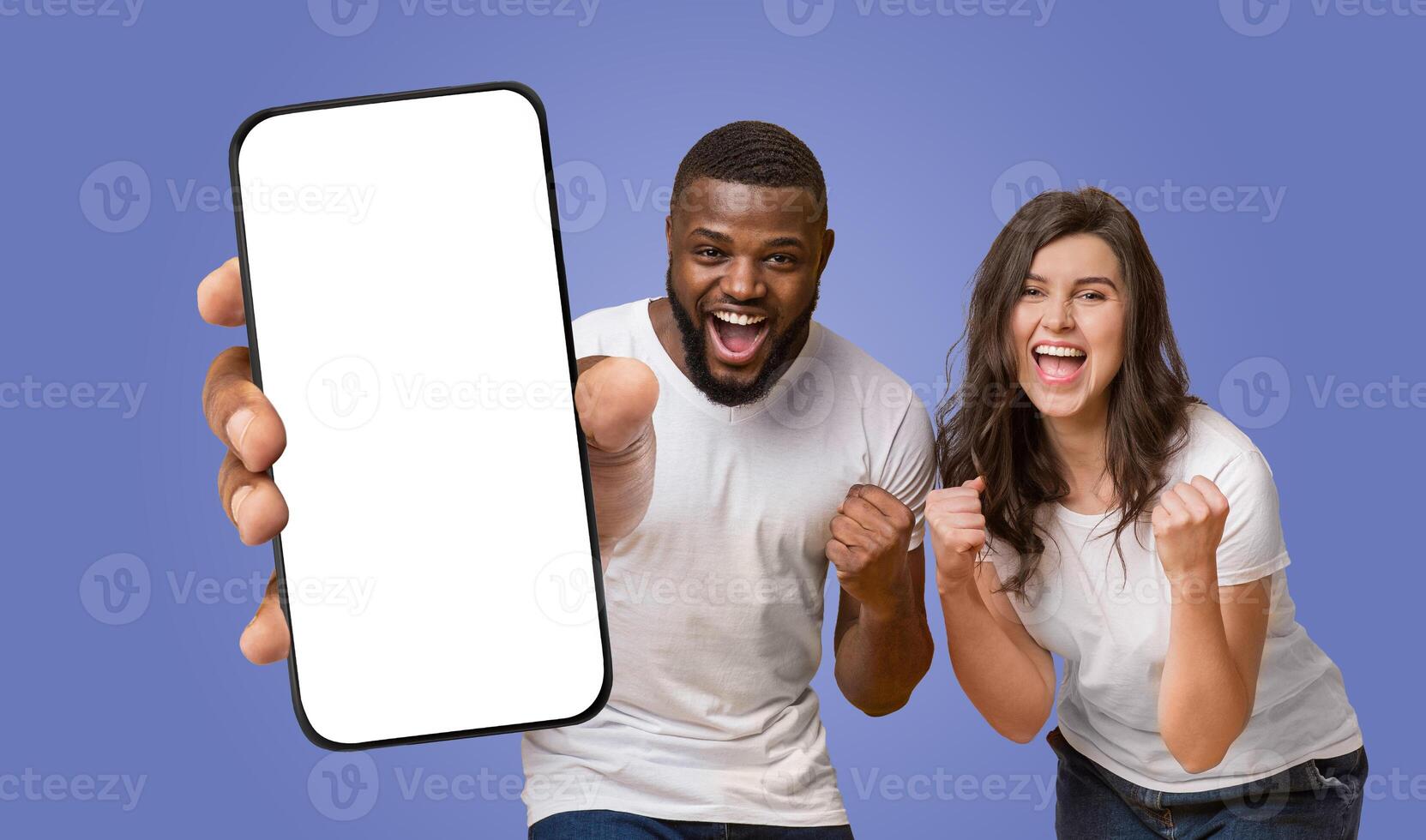 Multiracial couple standing in front of a purple background. The man is holding a smartphone with a blank screen towards the camera, while the woman is smiling and clenching fists photo