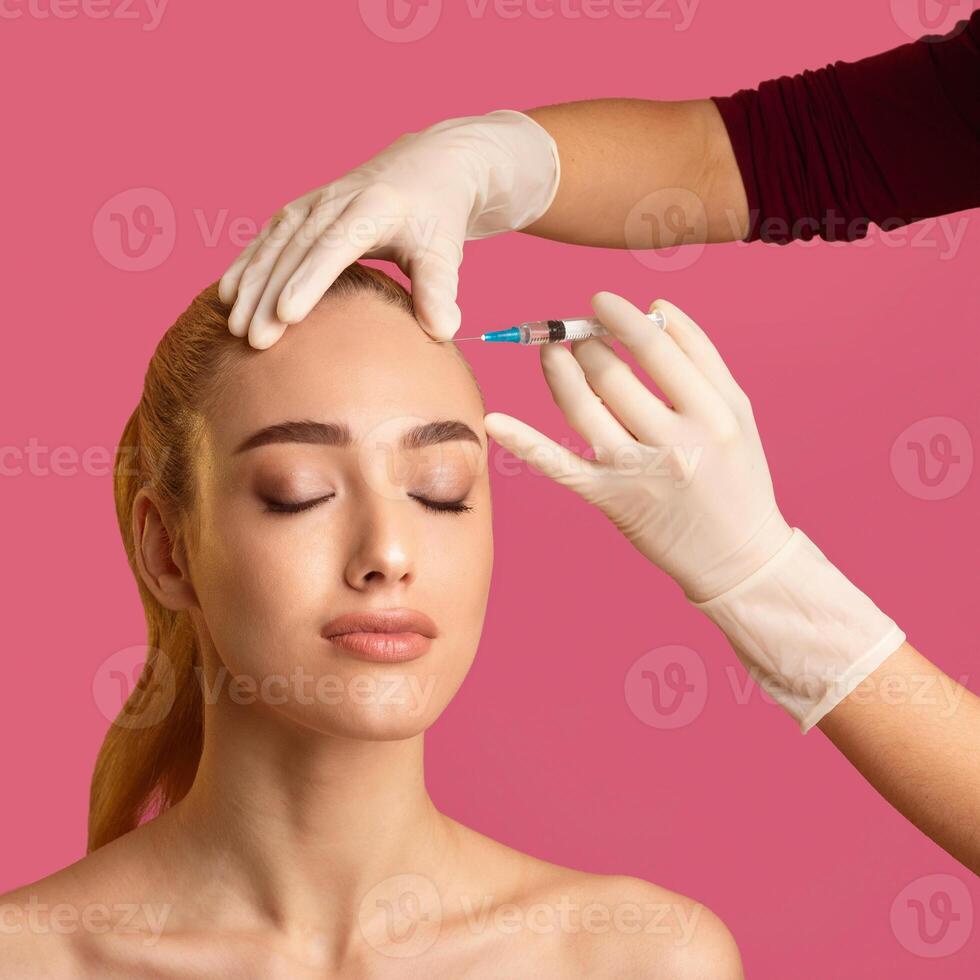 This image shows a young woman with blonde hair receiving a forehead injection treatment from a medical professional wearing gloves. The woman has her eyes closed photo
