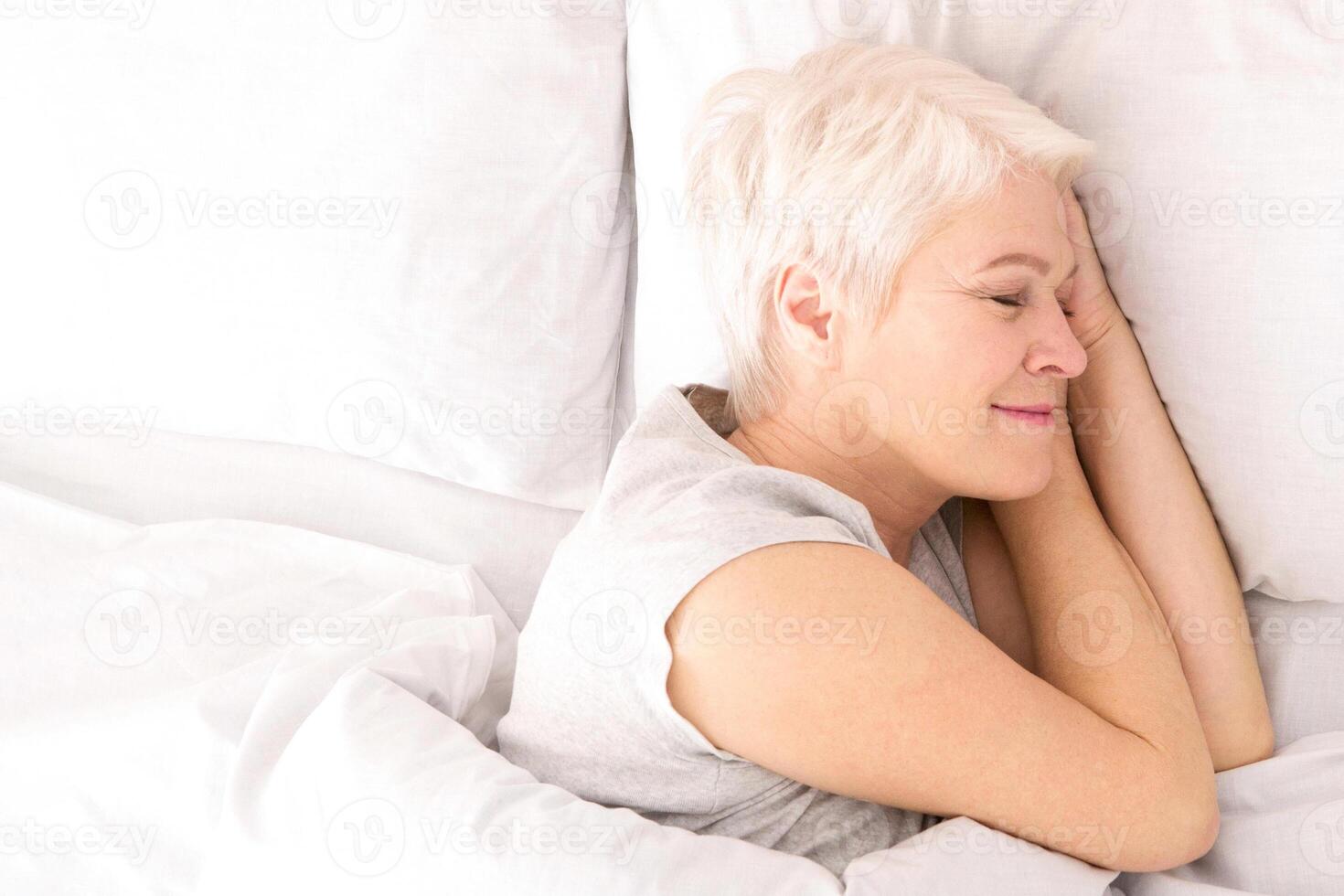 A woman is lying down in bed, covered by crisp white sheets. She appears relaxed and comfortable, with her eyes closed, possibly resting or sleeping, view above photo