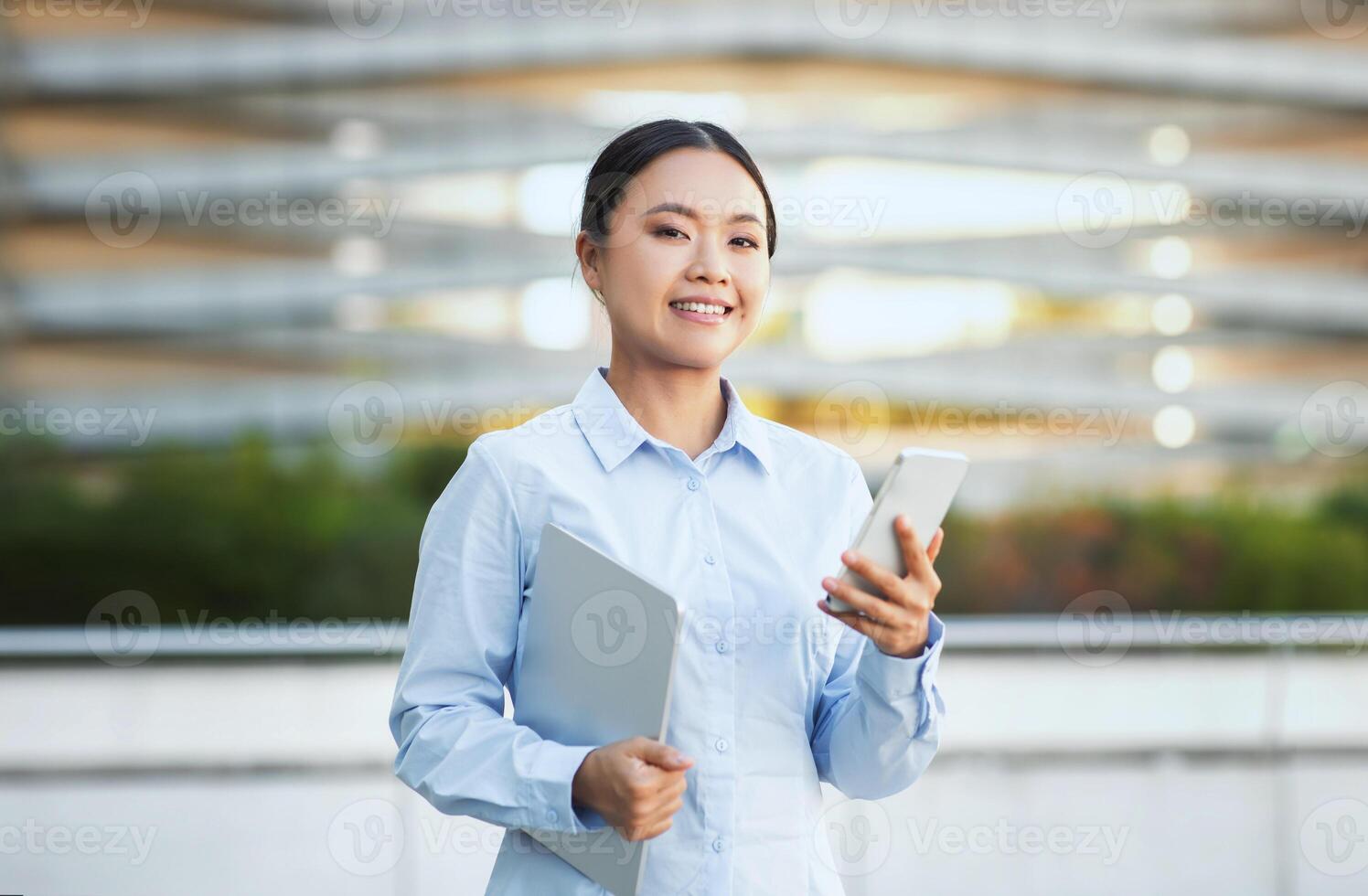 An Asian businesswoman is smiling while holding a smartphone in this stock . She appears professional and engaged, showcasing modern business practices. photo