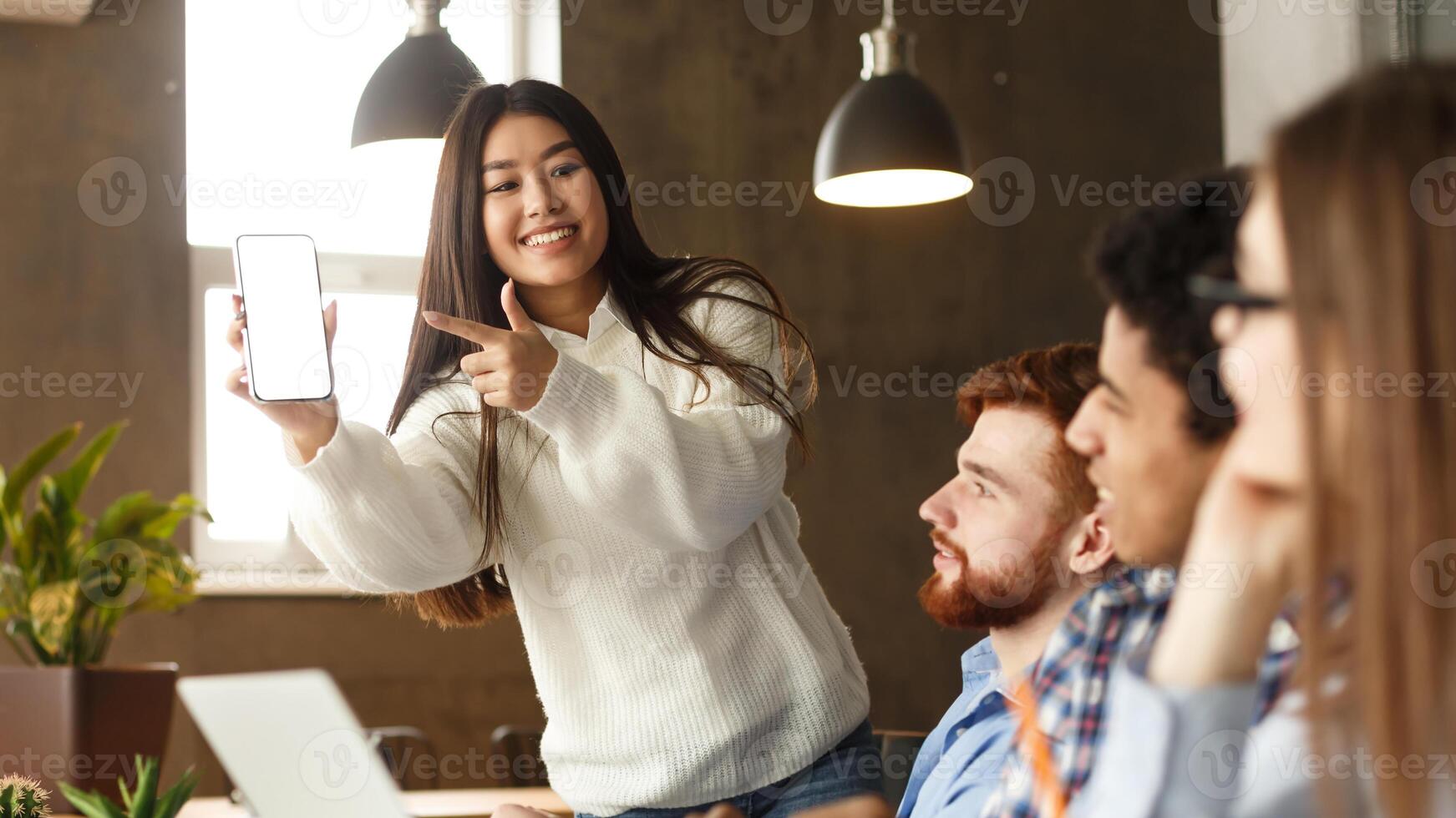 Asian girl showing cellphone to classmates, pointing at blank phone screen in library, free space photo
