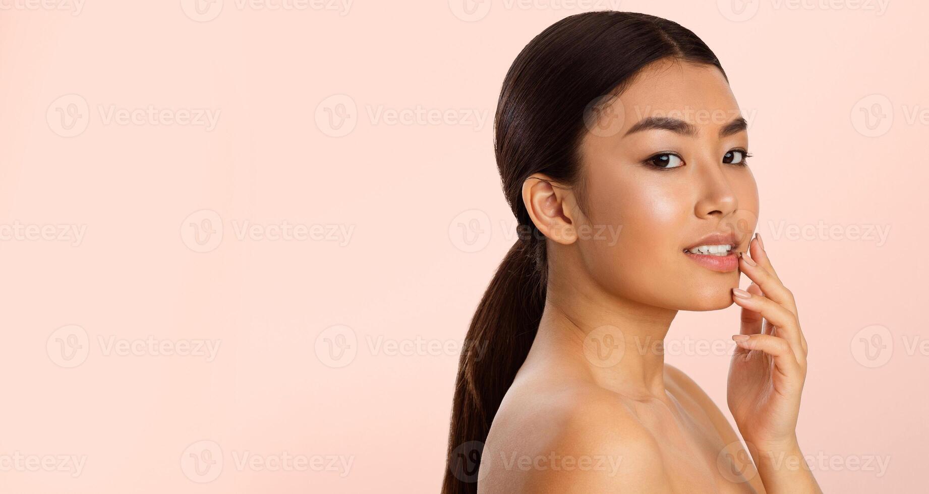 Asian woman with long dark hair in a ponytail smiles and touches her face gently. Her skin is clear and glowing and she is looking to the side. The background is a soft, light pink. photo