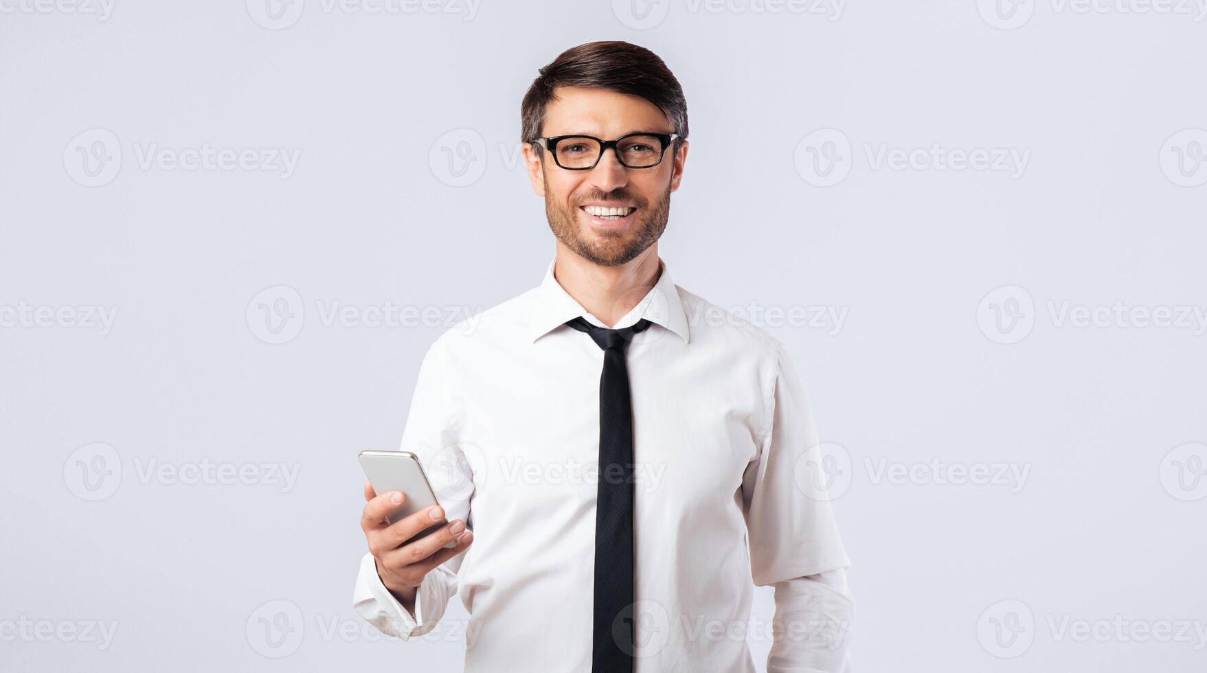 A man in a white shirt and tie smiles while holding a smartphone. He is standing in front of a white background. photo