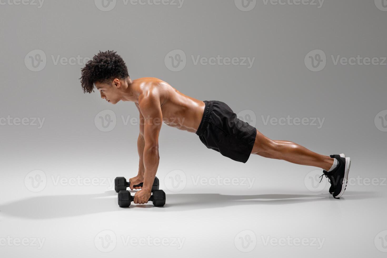 A fit young man engages in a rigorous workout by executing dumbbell push-ups in a well-lit studio gym. He maintains proper form while focusing on strength training and building muscle endurance photo