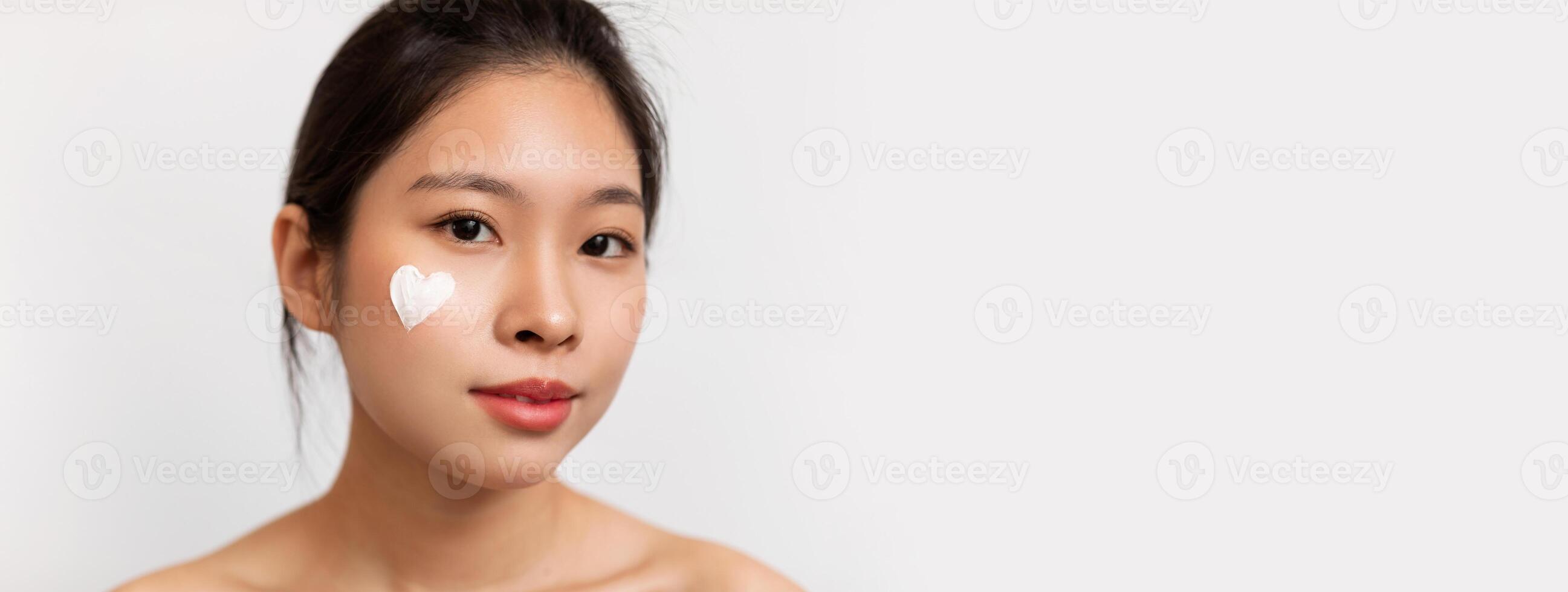 Closeup portrait of pretty korean lady with moisturizer cream on her face in heart shape, woman testing new beauty product, white studio background. Face care concept, copy space photo