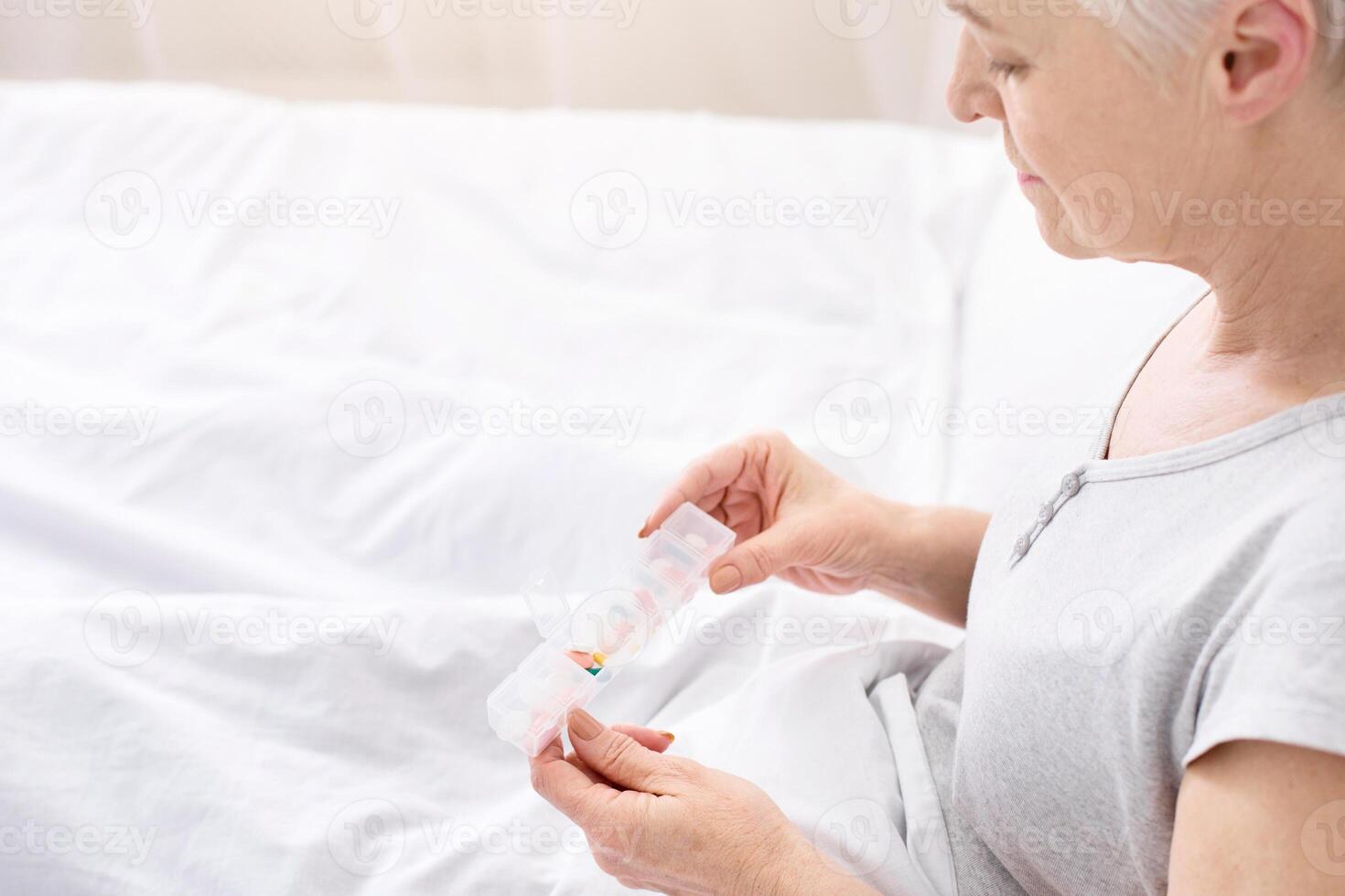 An elderly woman with short hair is sitting on her bed, carefully sorting an assortment of colorful pills and capsules into a daily pill organizer, indicating a morning medication management routine. photo