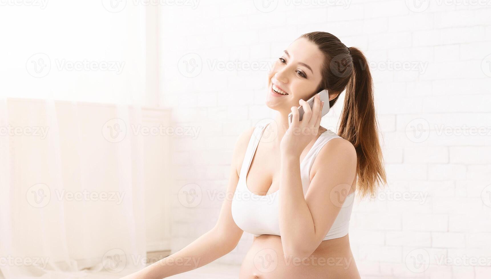 A pregnant woman is happily speaking on the phone while seated in a bright, airy room with white decor, wearing casual, light-colored clothing and appears to be in a positive mood, smiling widely. photo