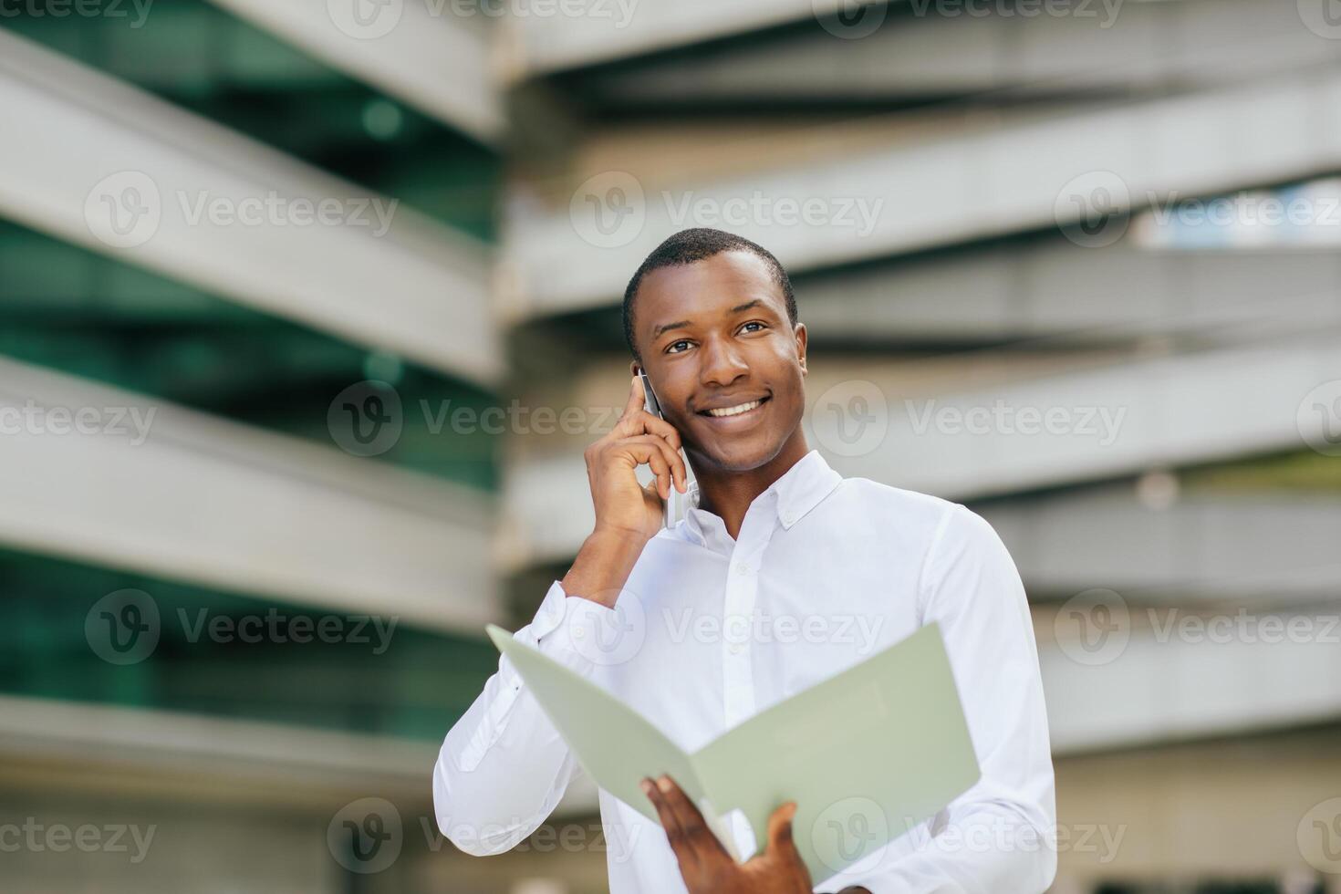 Black man in a white shirt holds a folder in his left hand and talks on his phone with his right hand. He is standing in front of a large building with a modern design photo