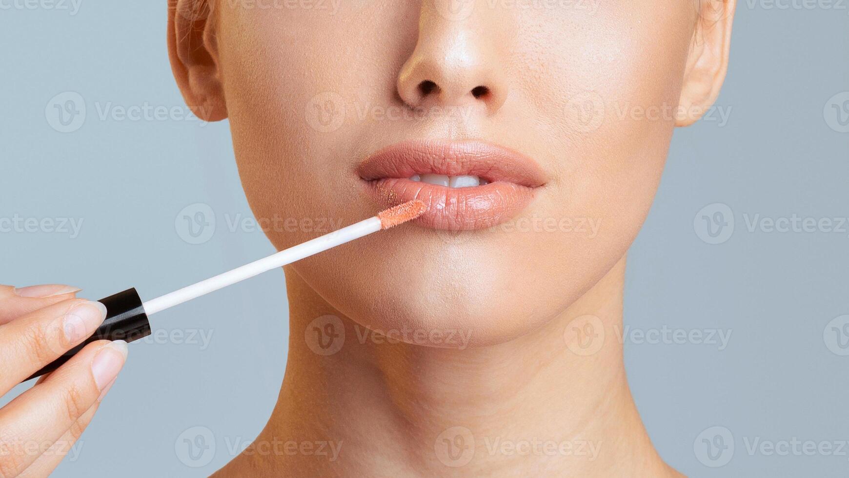 A woman with smooth skin is applying lip gloss to her lips with a clear applicator wand. The background is a light blue, cropped photo