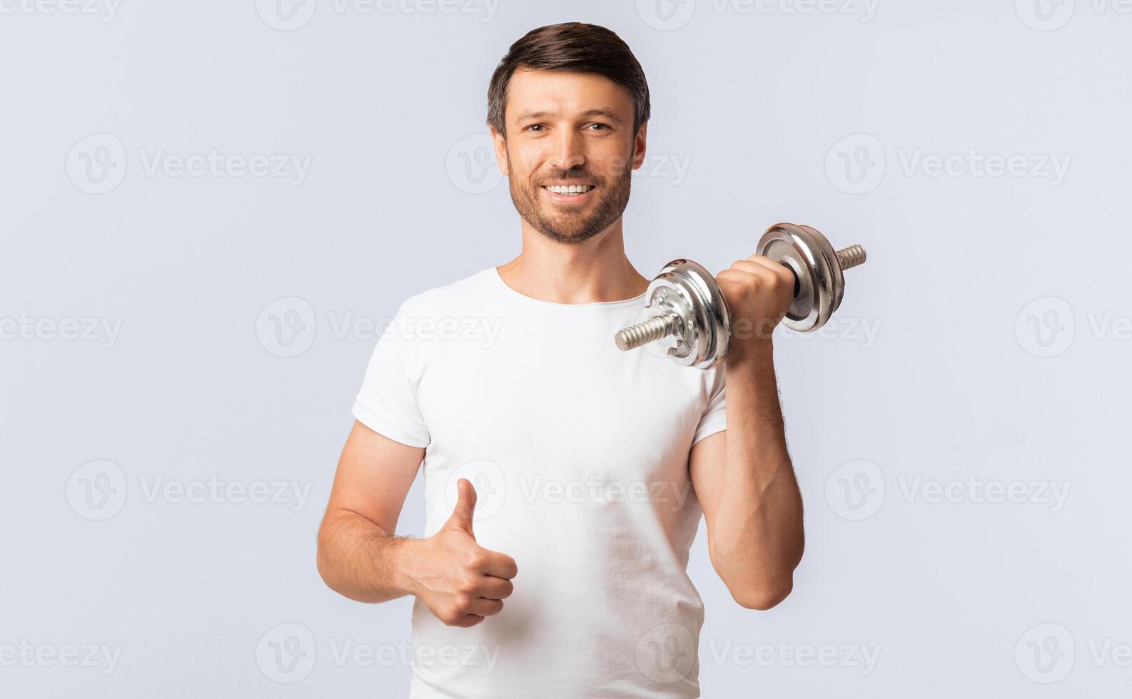 A man in a white t-shirt is lifting a dumbbell with his right arm and giving a thumbs up with his left hand. He is smiling and looks happy and healthy. photo