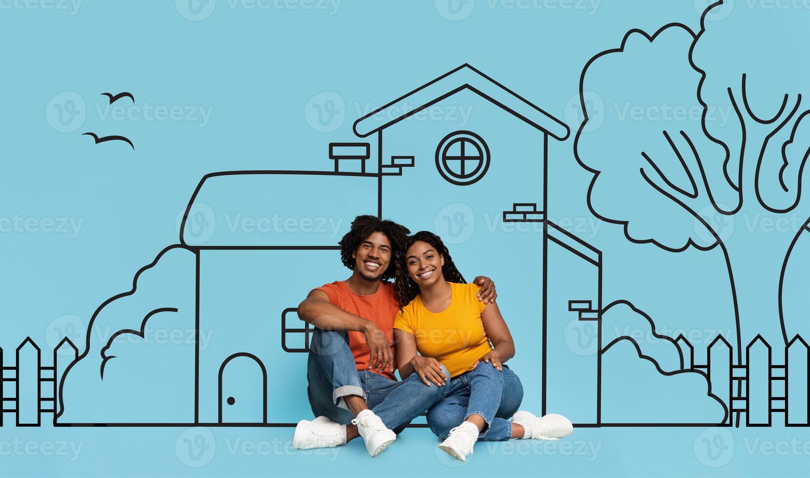 A cheerful young black couple is sitting closely together against a backdrop with a drawing of a house, trees, and birds. The drawing implies dream or goal of homeownership, or simple life together photo